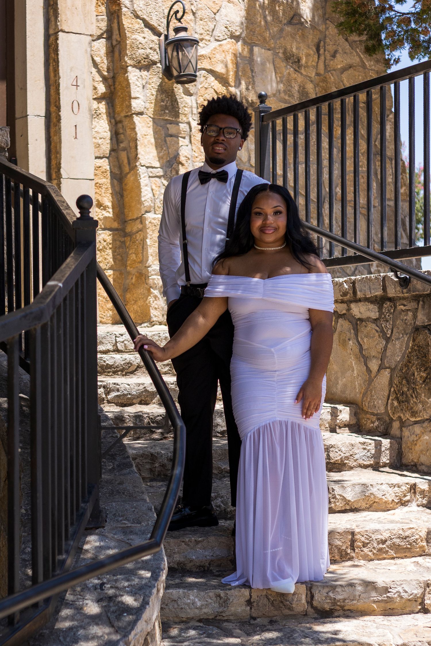 A young couple dressed in formal prom attire poses on stone steps beside black iron railings.