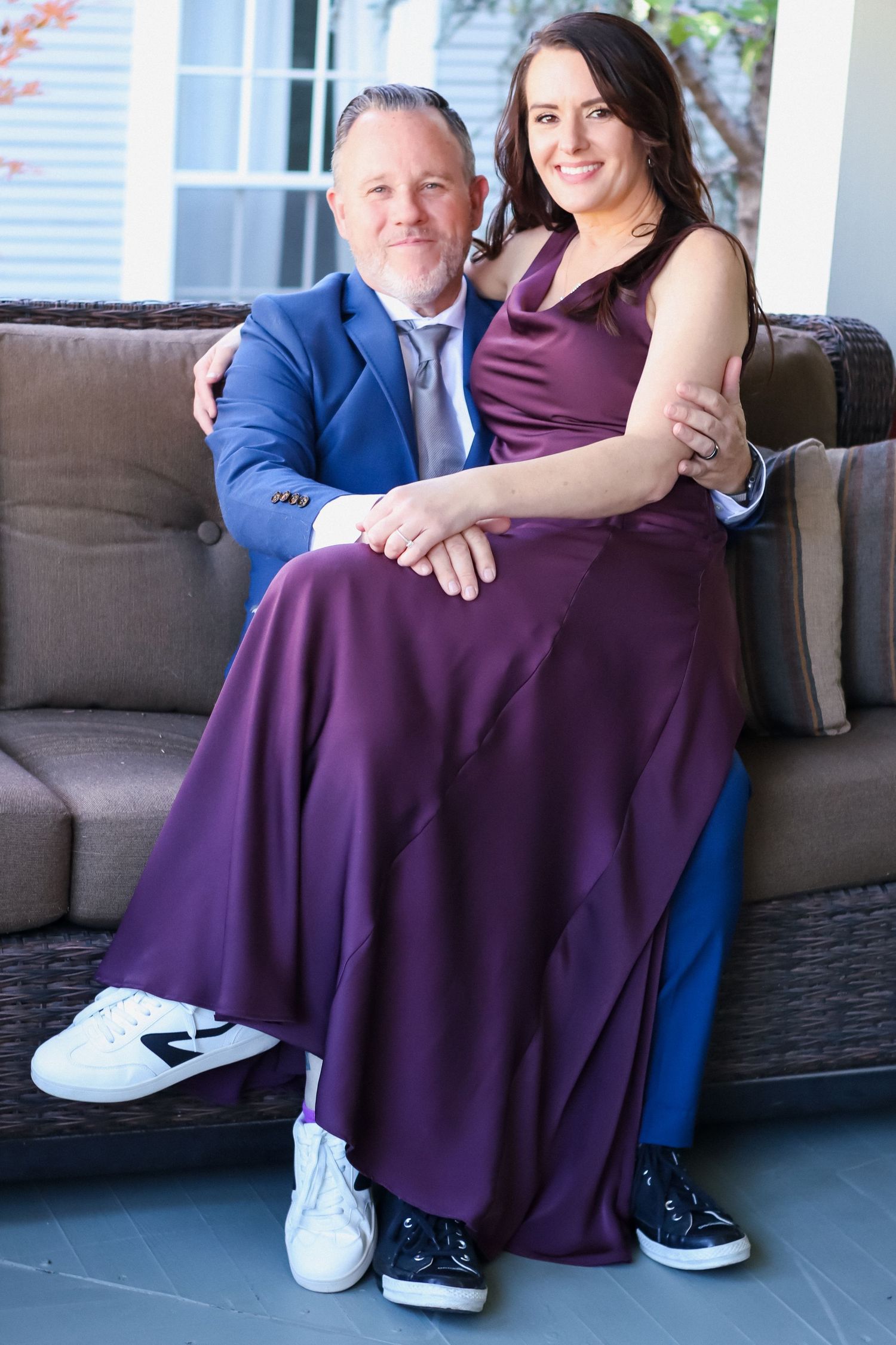 A couple in formal attire sits together on a grey outdoor couch wearing matching sneakers.