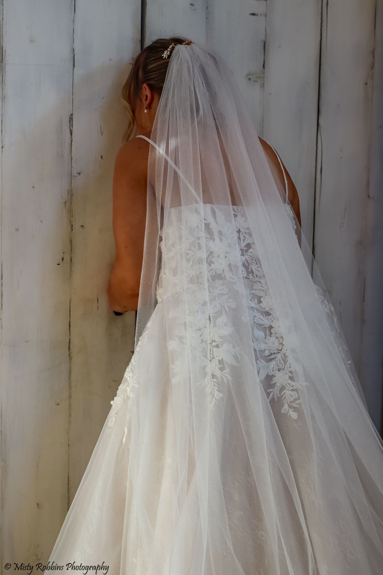 Back view of a bride in a flowing white wedding gown with delicate lace details and sheer veil against white wall.