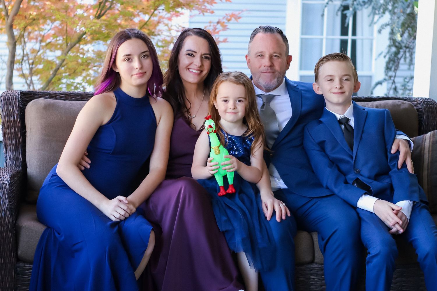 Family of five posing together on a porch, dressed in formal blue and purple attire, with autumn trees in the background.