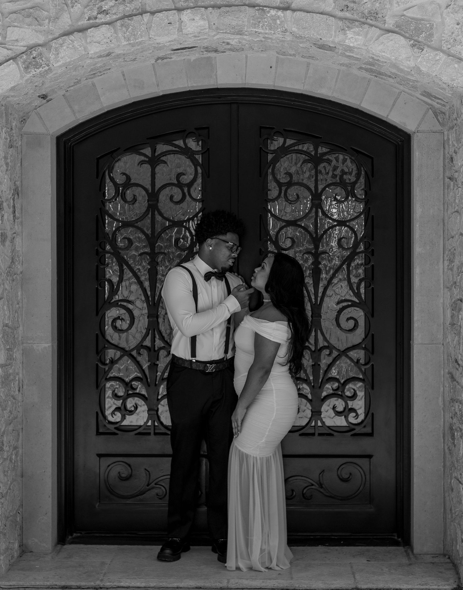 Black and white photo of a couple in formal wear sharing a romantic moment before ornate iron doors.