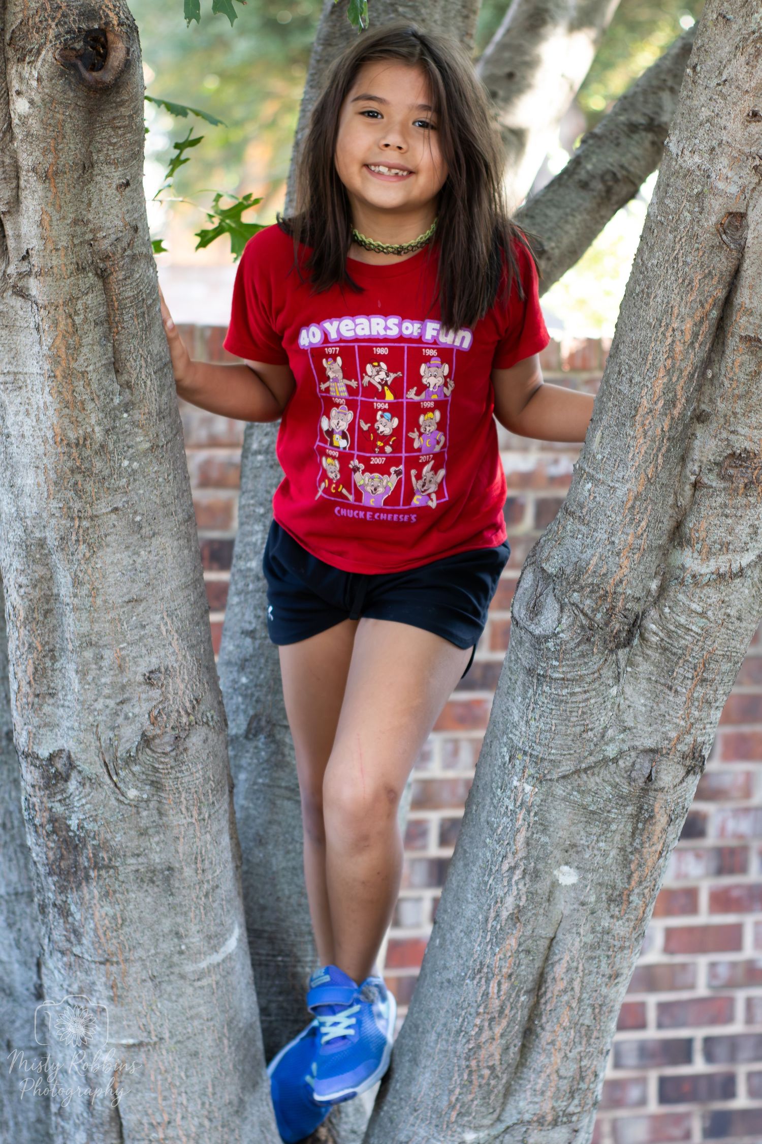 Child in red t-shirt climbs tree branches against brick wall background in playful outdoor scene.