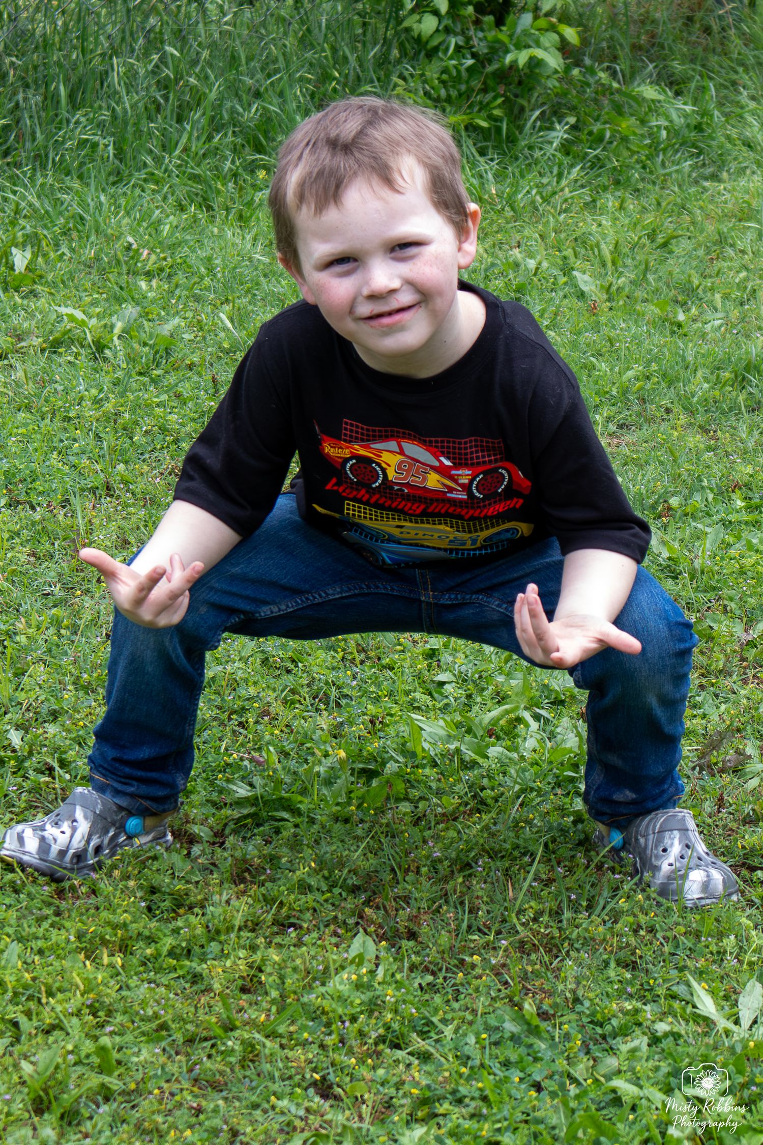 Young boy in a Cars movie shirt squatting on green grass, smiling and posing with hands outstretched.