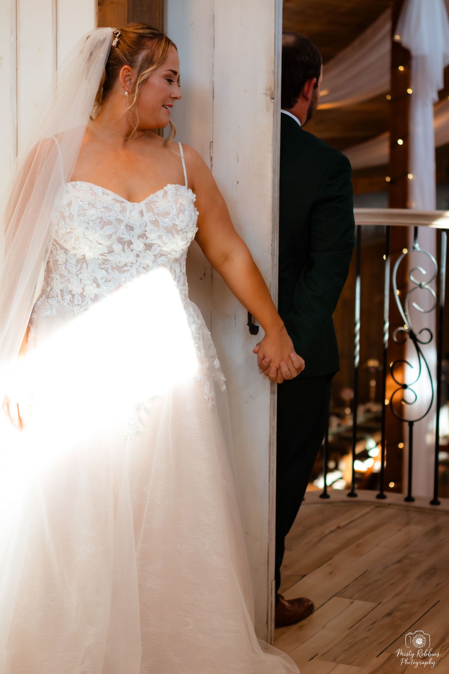 A bride in a white wedding gown with lace details stands gracefully against a rustic wooden wall.
