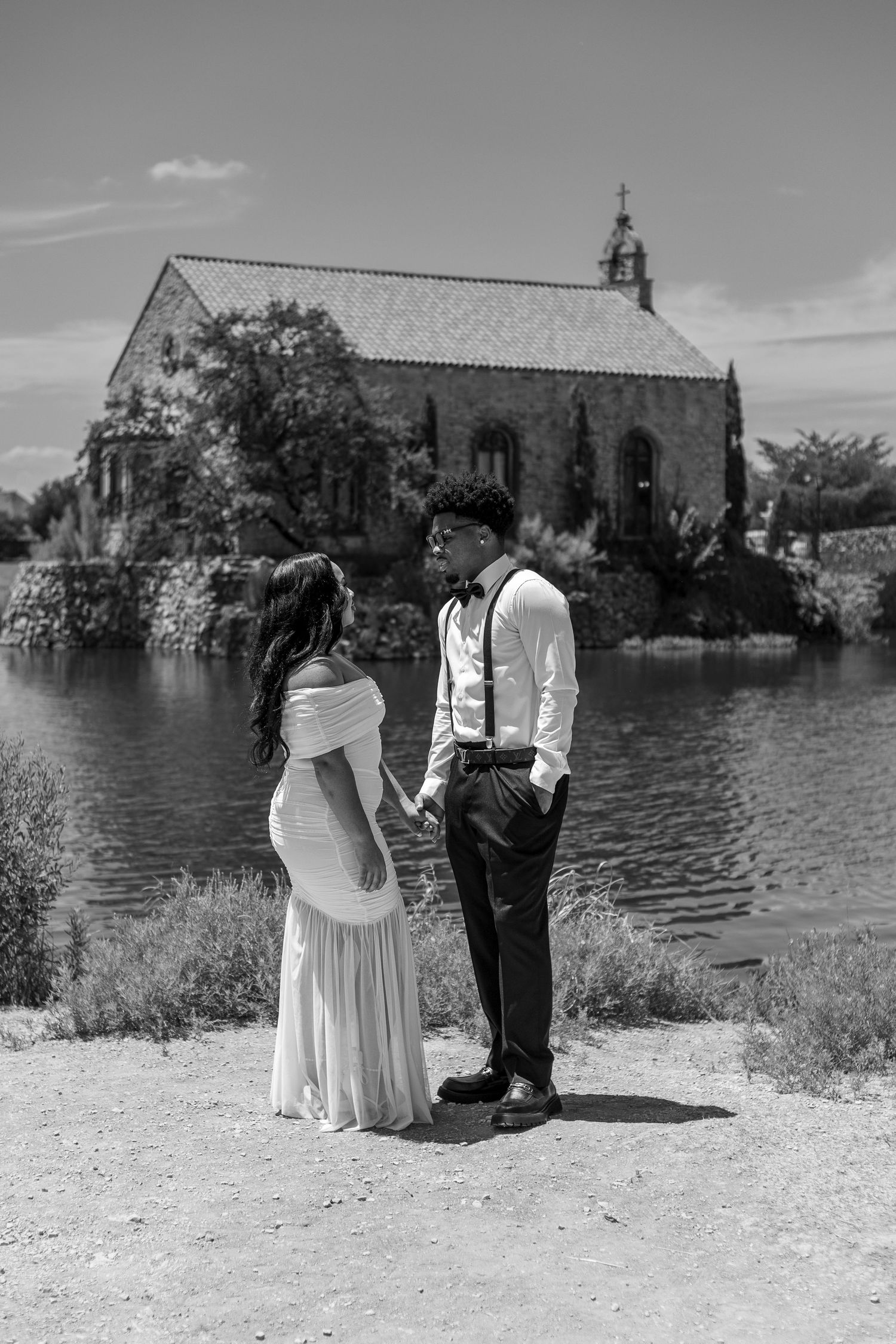 Black and white photo of a couple holding hands near a lake with a historic stone chapel in the background.