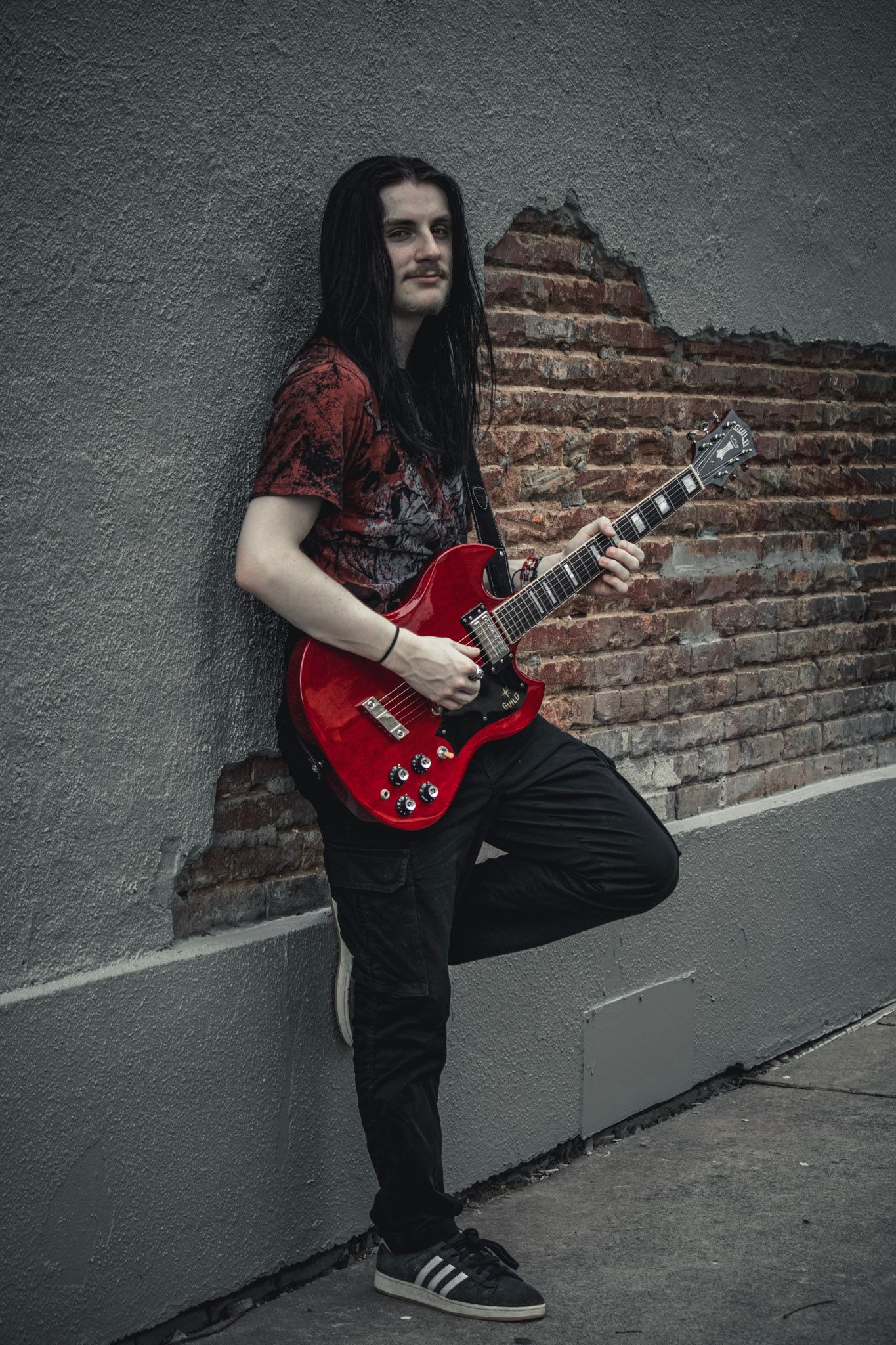 A musician in black pants and red shirt plays an electric guitar against a brick wall in an urban setting.