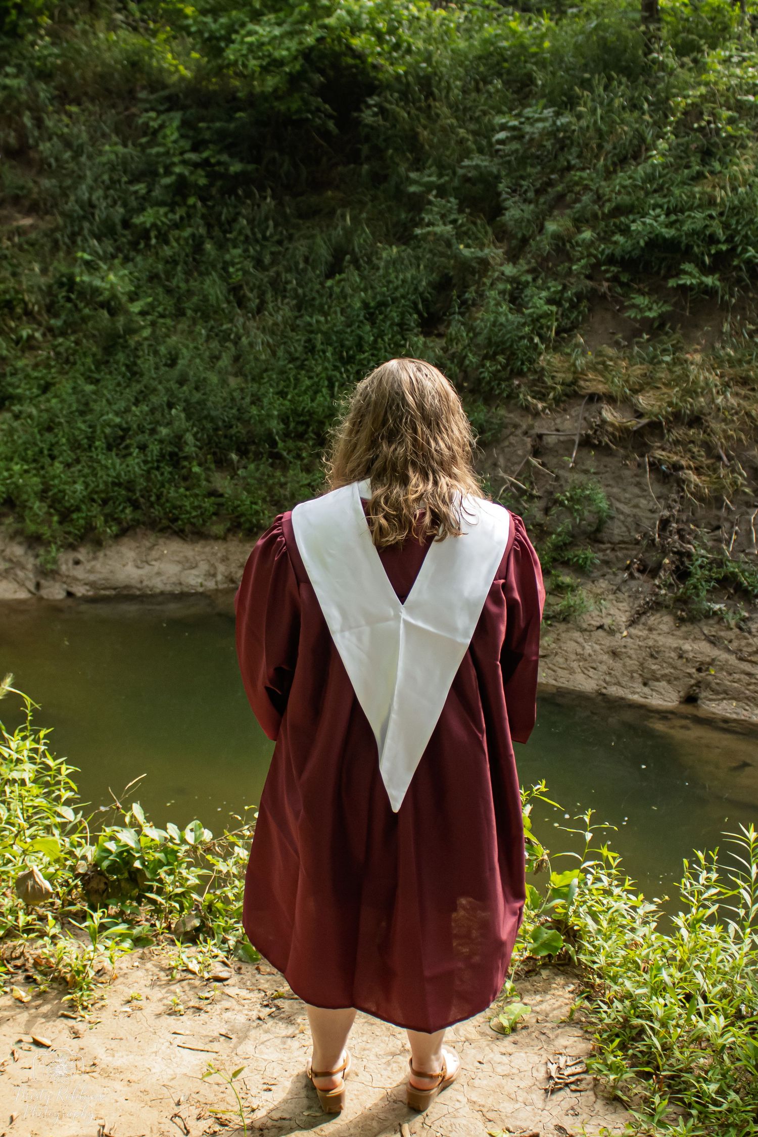 A person in a burgundy graduation gown with white collar stands at the edge of a pond surrounded by greenery.