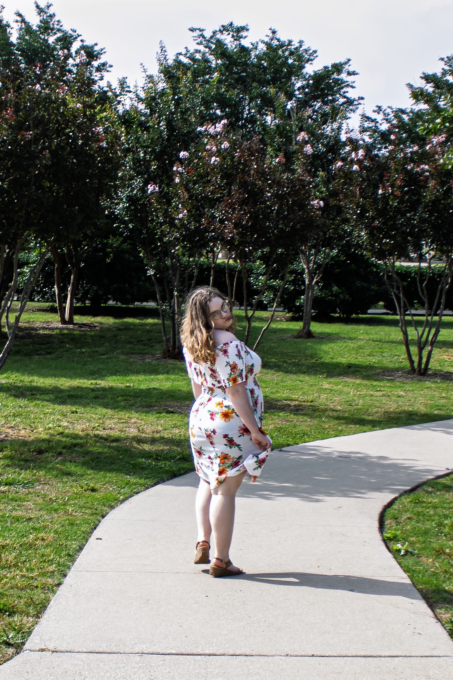 A person in a butterfly print dress walks along a winding concrete path through a green park setting.