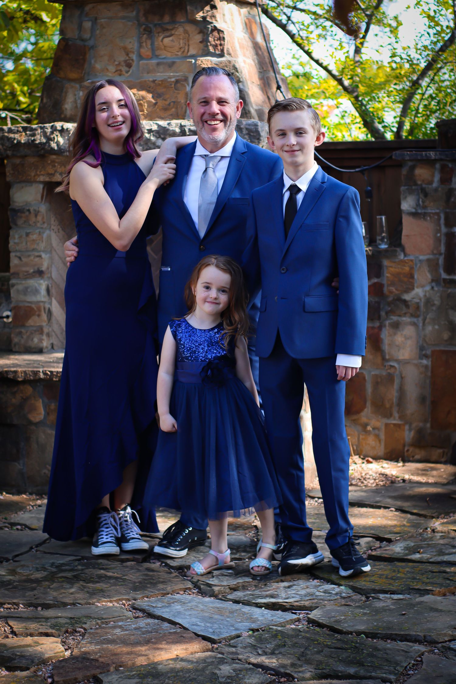 A family of four poses together in coordinating formal navy blue attire against a stone wall backdrop.