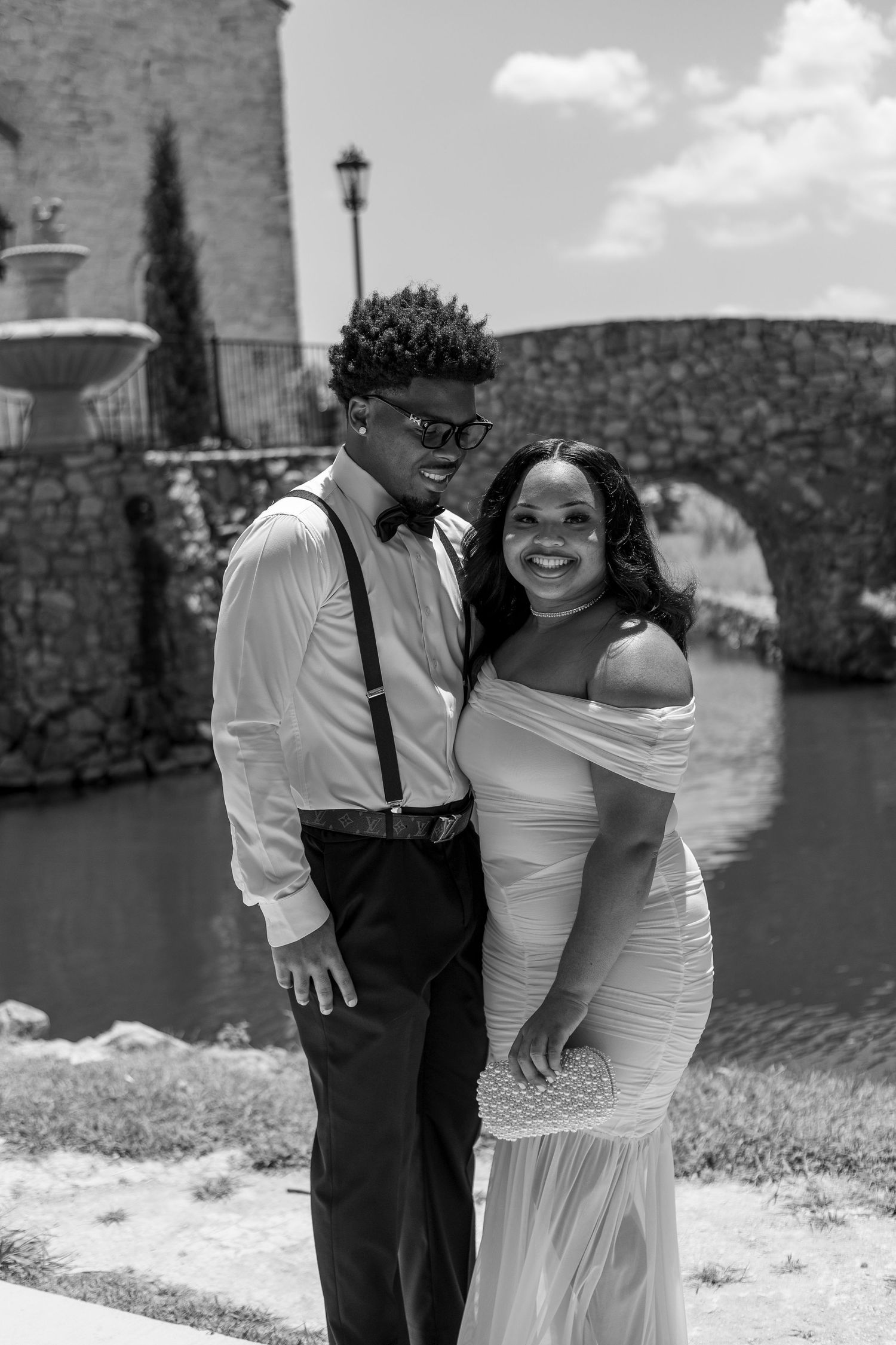 Black and white photo of a smiling couple posing outdoors near a stone bridge and fountain, man in suspenders and bow tie.