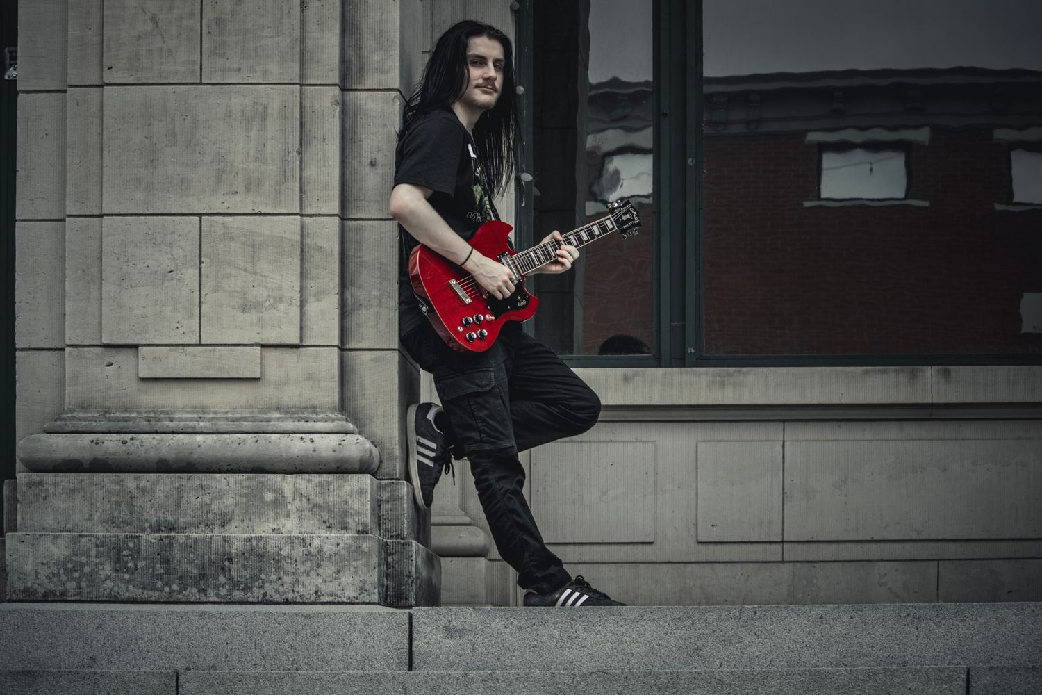 Person in dark clothing playing red electric guitar while leaning against stone pillar.