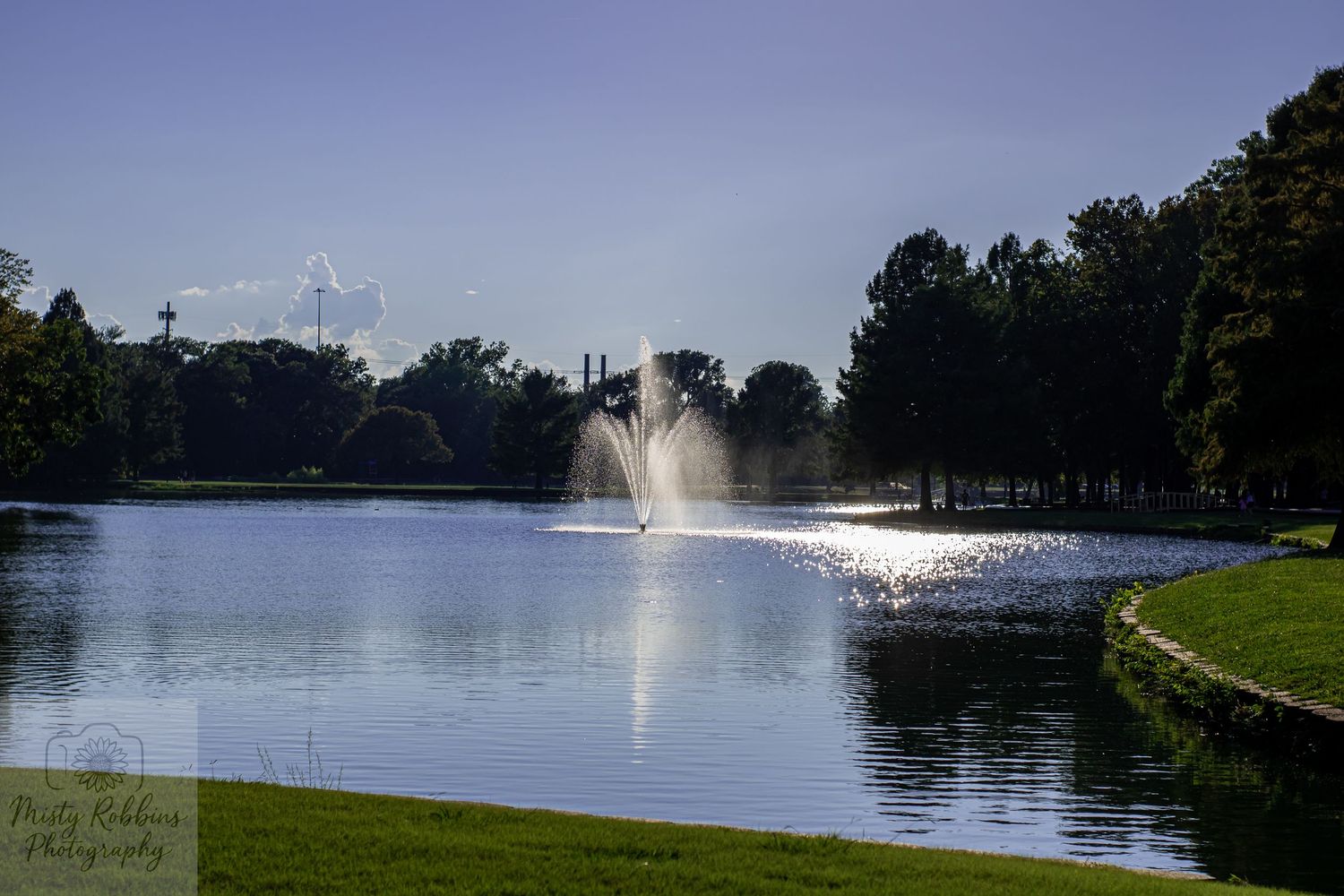 Panoramic view of a serene lake with fountains spraying water against a bright sky.