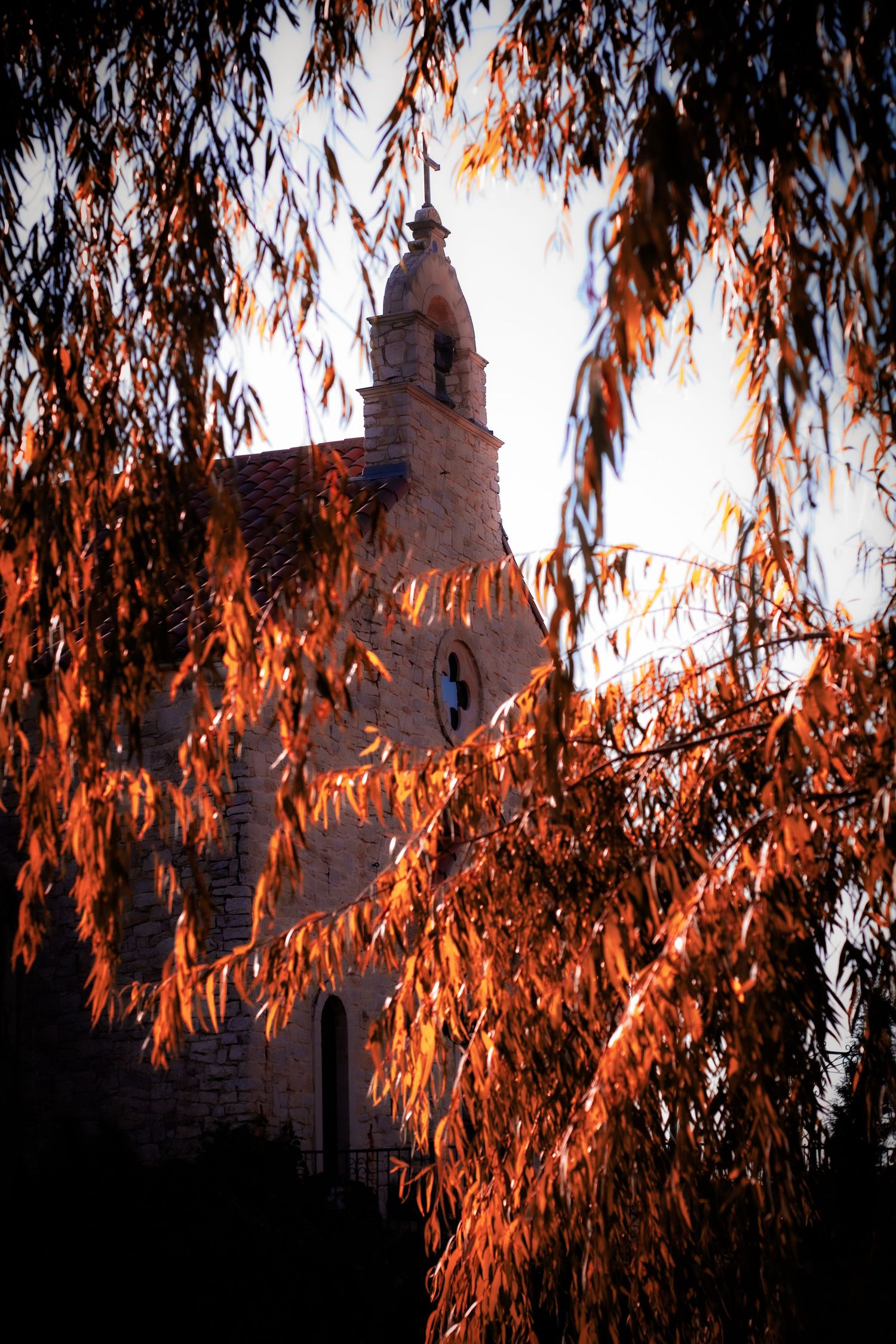 Historic stone church with bell tower framed by autumn orange foliage branches against a bright sky.