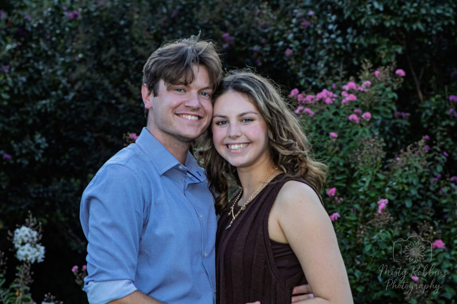 A couple poses together in a garden setting with pink flowers blooming in the background.