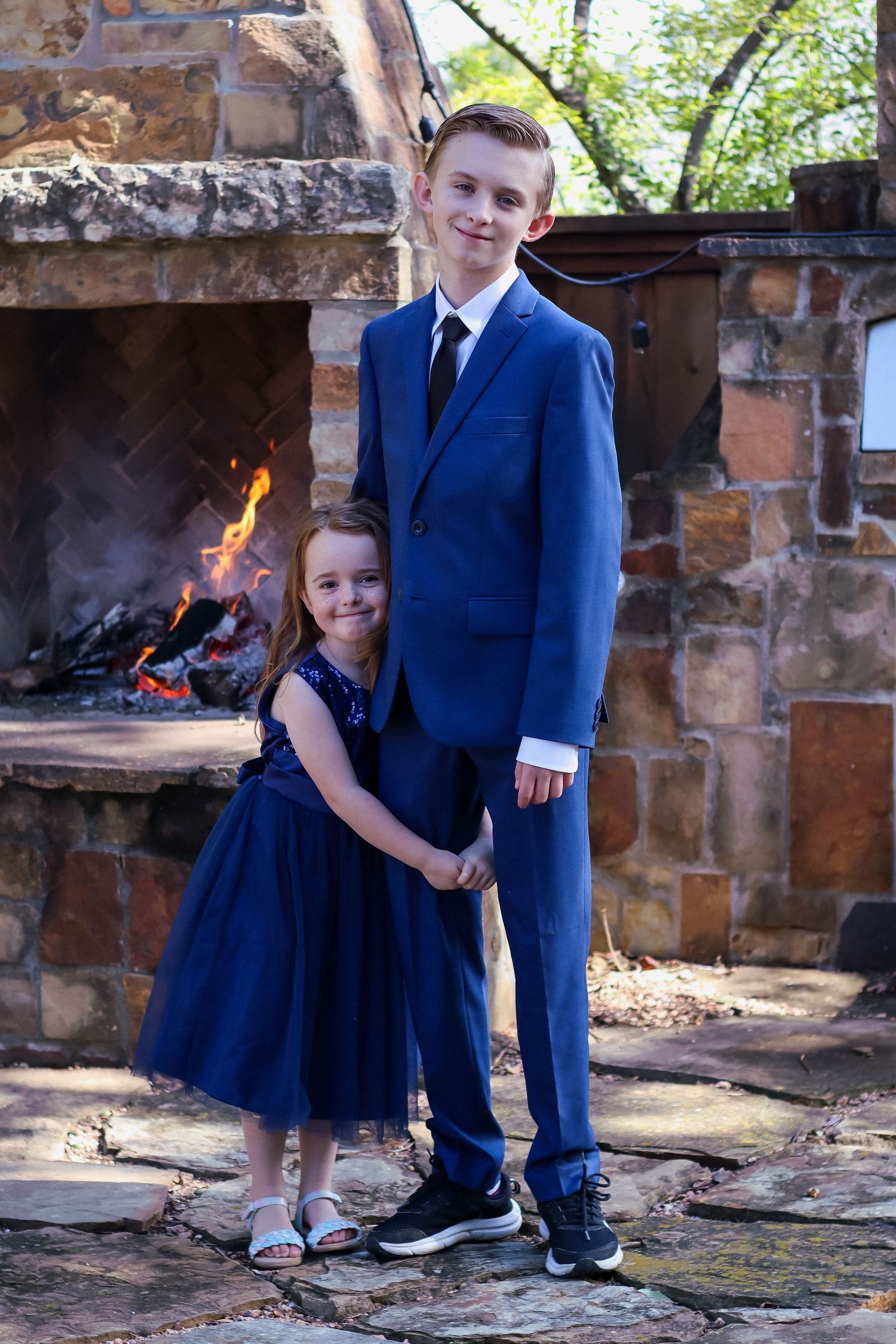 Two people in coordinating navy formal wear pose in front of an outdoor stone fireplace.