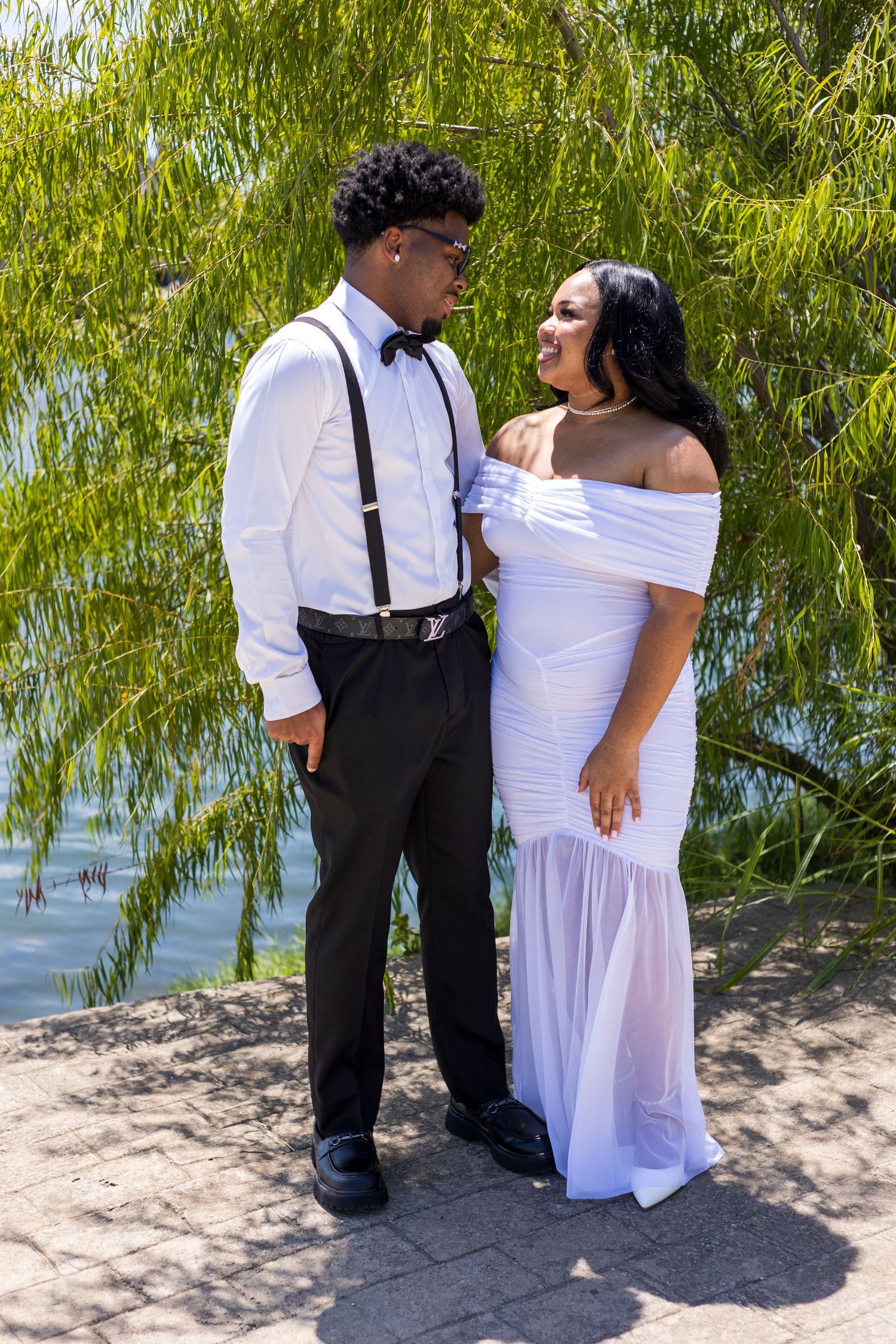 A young couple in formal attire poses outdoors near a willow tree, wearing white dress and black tuxedo.