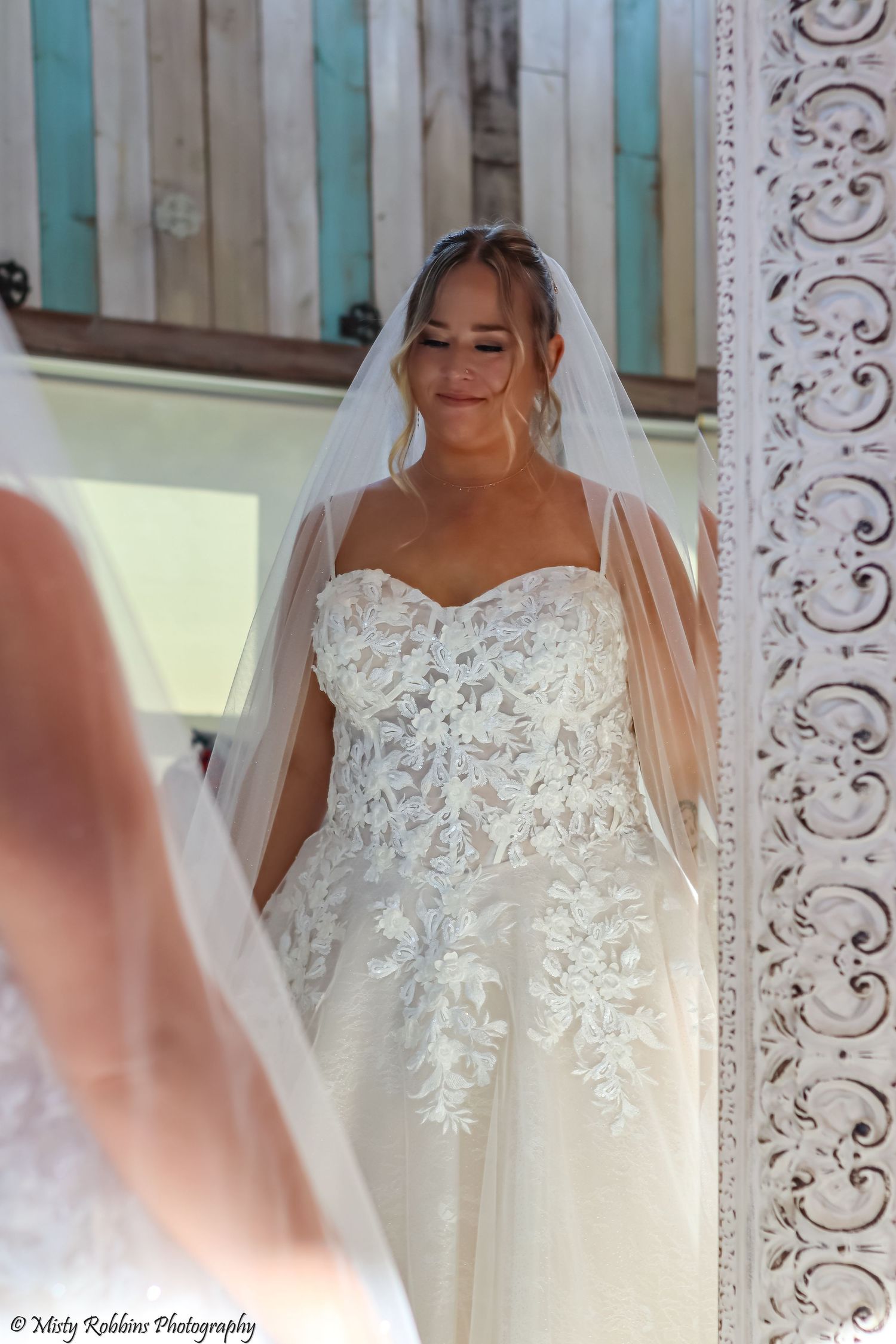 Bride in lace strapless wedding gown and veil smiling at her reflection in an ornate white mirror.