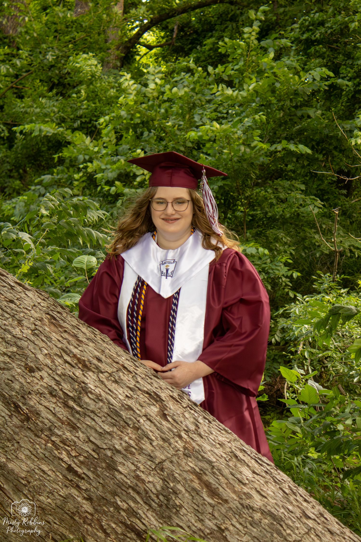Smiling graduate in maroon cap and gown poses outdoors against lush green foliage near a large tree trunk.