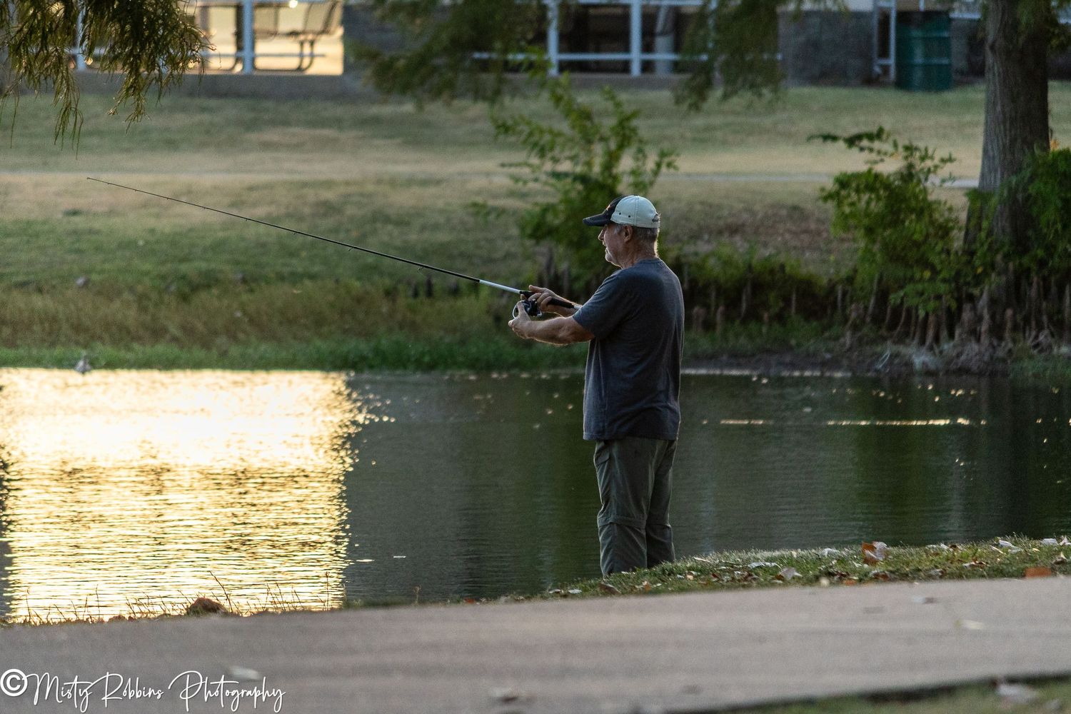 Man fishing at a calm pond during golden hour sunset in a park setting.