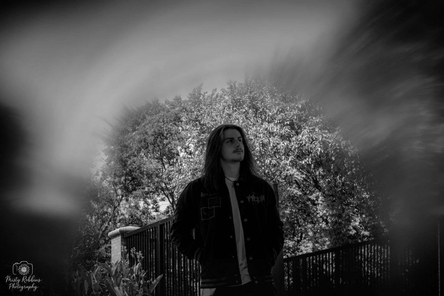 Black and white portrait of a long-haired person in a denim jacket standing outdoors near trees and a fence.