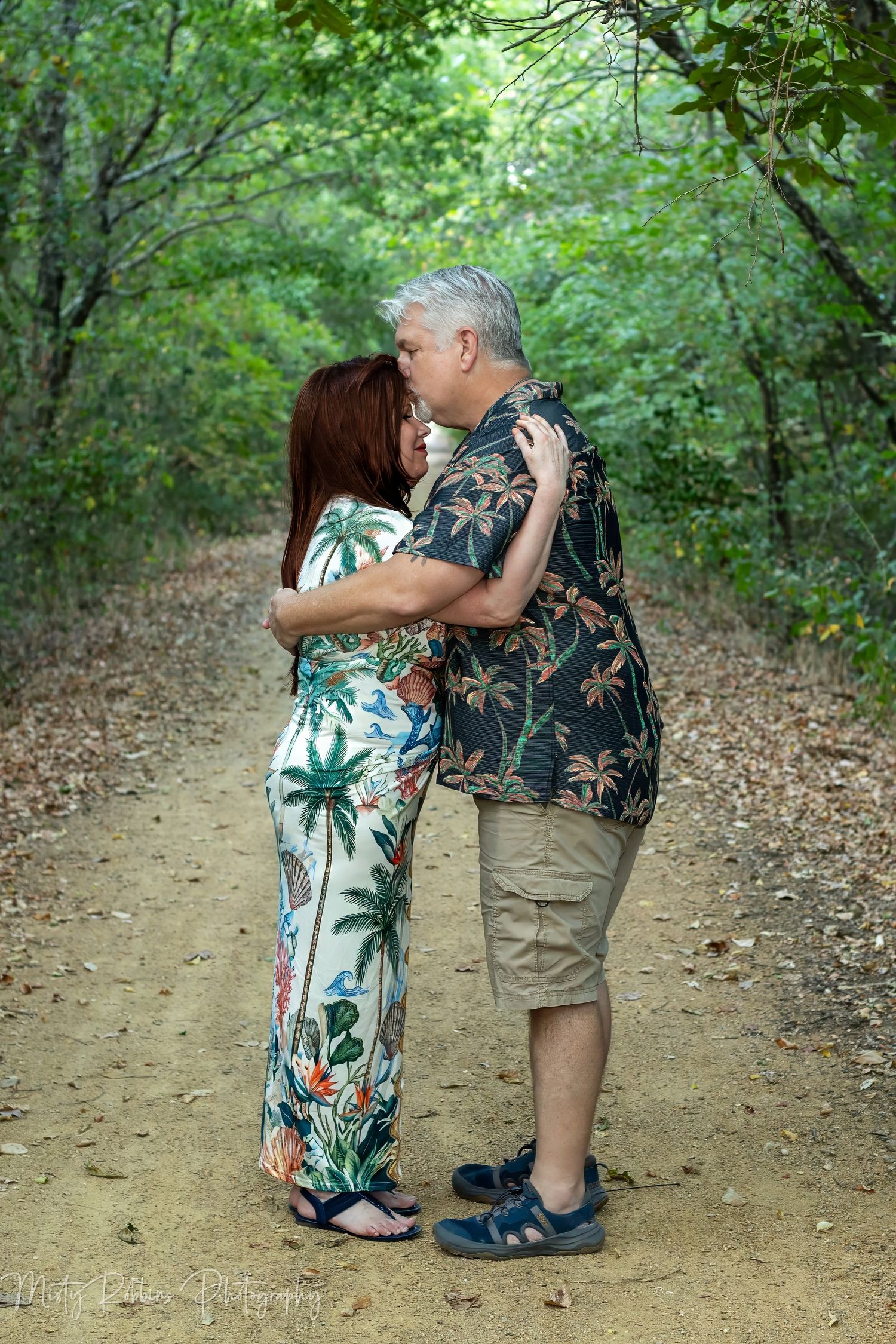 Couple embracing and kissing on a scenic forest path surrounded by lush green trees and fallen leaves.