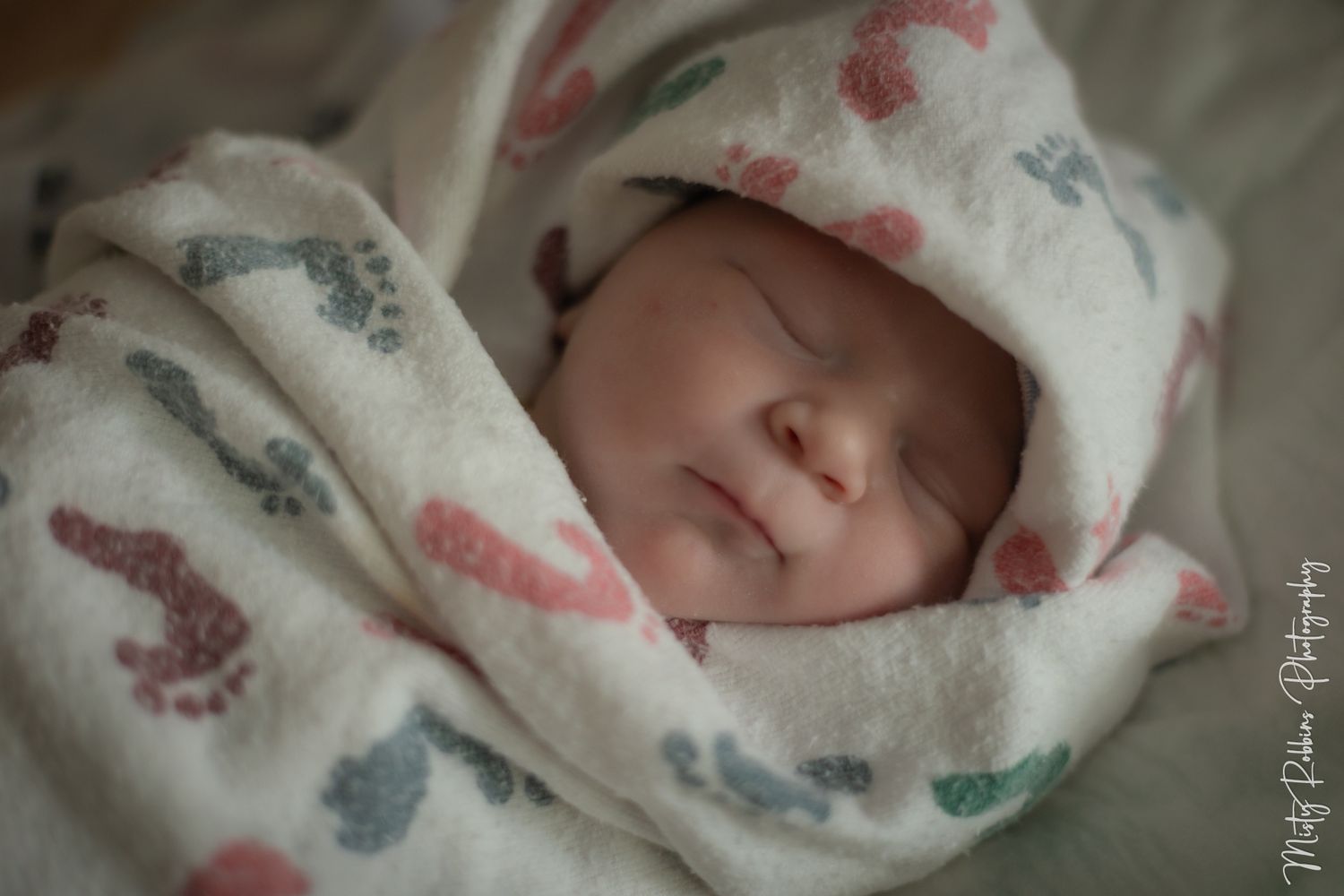 Newborn baby sleeping peacefully wrapped in a soft white blanket with colorful footprint pattern.
