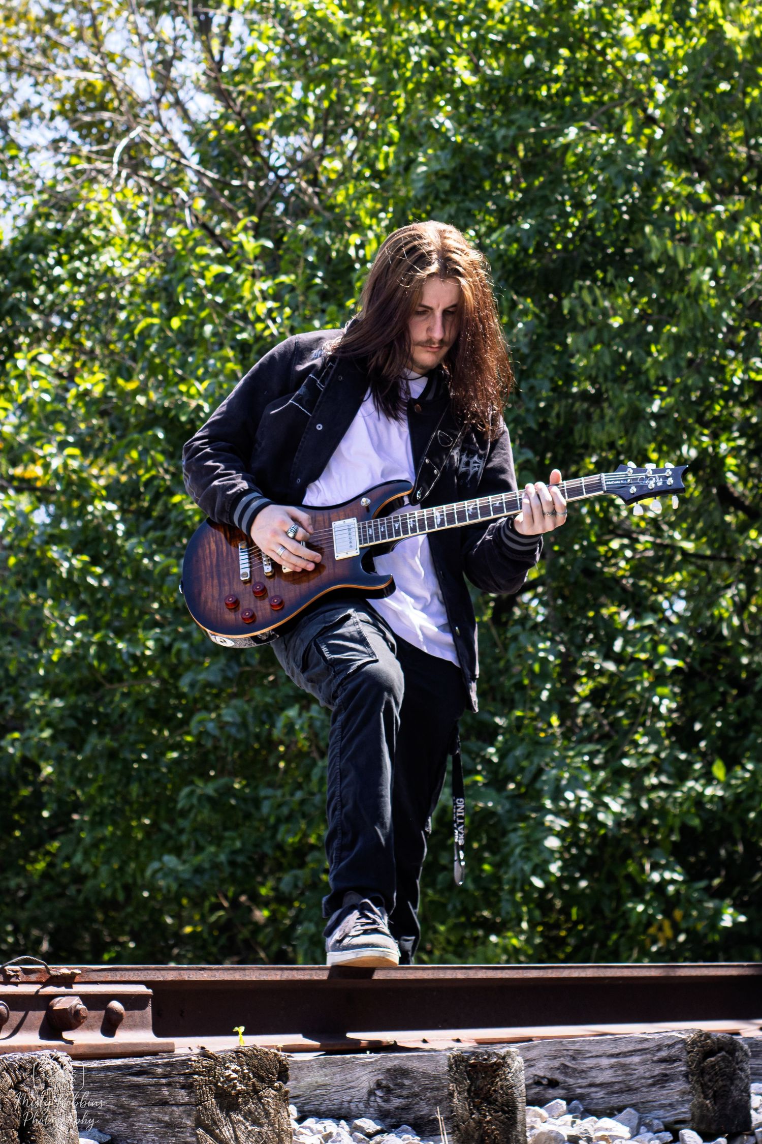 Musician in black leather jacket plays electric guitar while balancing on railroad tracks in outdoor setting with green foliage.