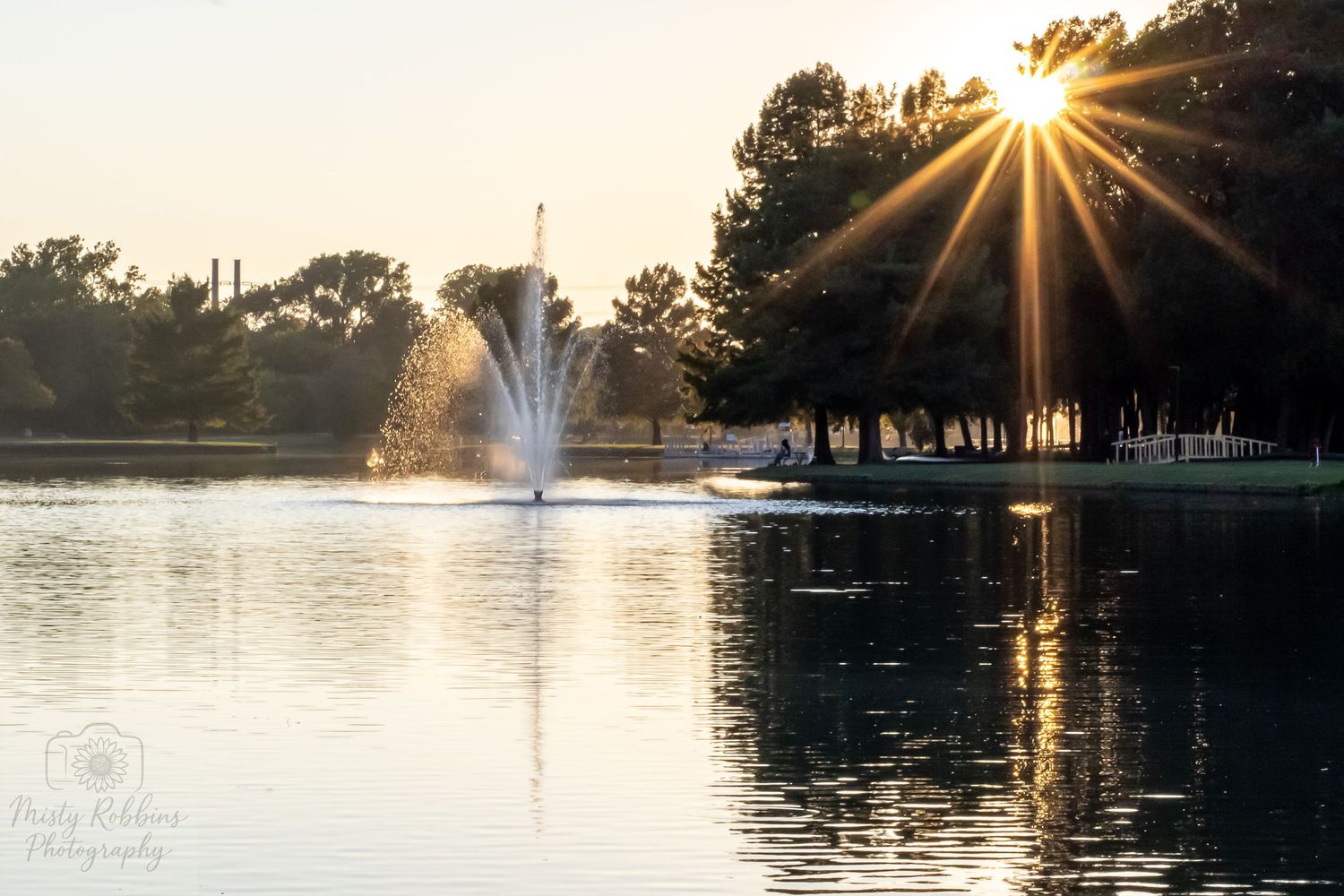 A serene lake at sunset with water fountains creating a misty display as golden sunbeams shine through trees.