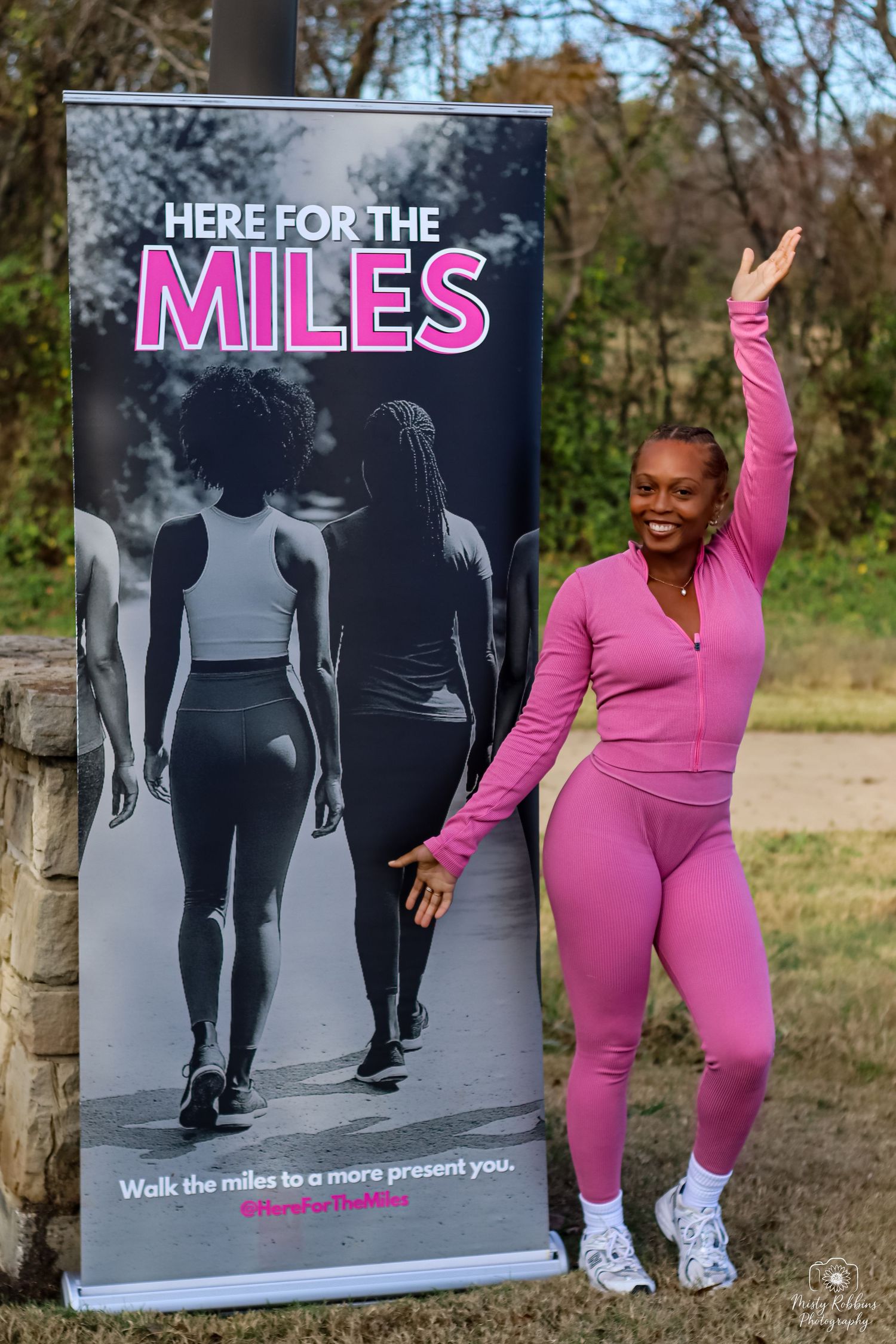 Woman in pink athletic wear poses beside a Here For The Miles fitness banner in an outdoor park setting.