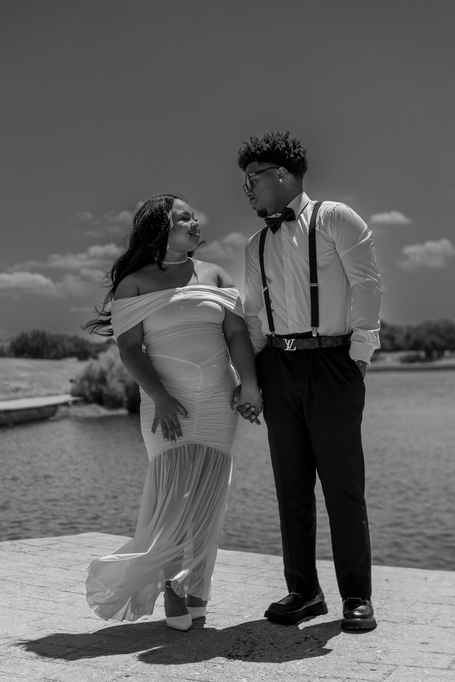 Black and white photo of a couple in formal wear holding hands by a lake, woman in white gown, man in suspenders.