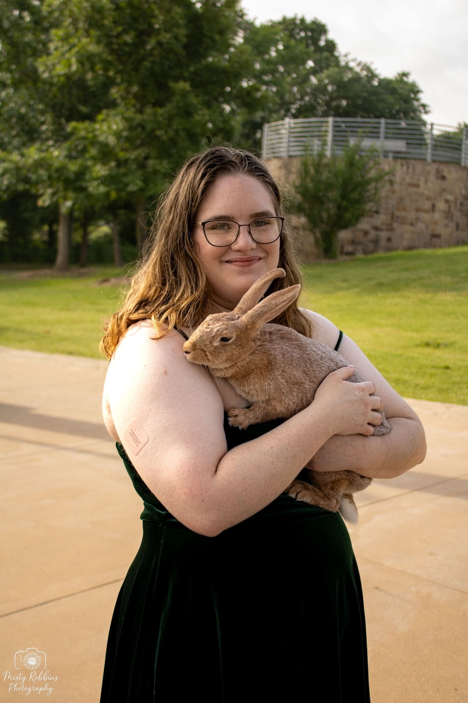 A person in a black dress holds and cuddles with a brown rabbit outdoors on a sunny day.