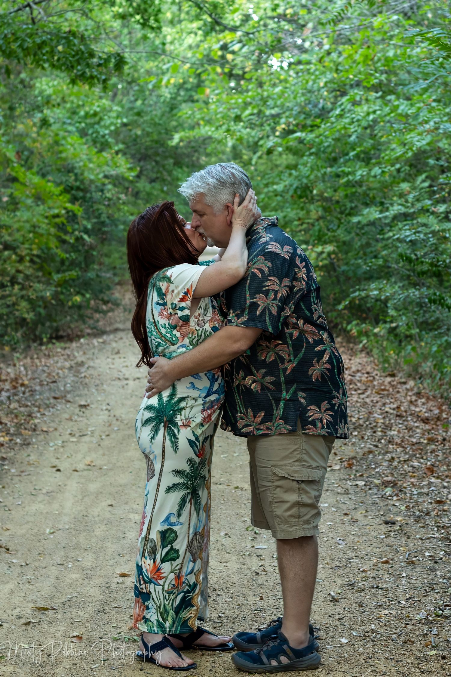 Couple sharing a romantic kiss on a wooded dirt path, wearing tropical print outfits, surrounded by lush green trees.