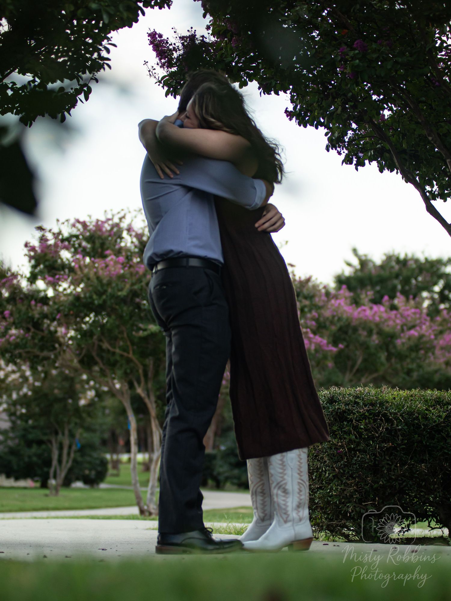 A couple embraces in a romantic outdoor setting with lush trees and pink flowering bushes in the background.