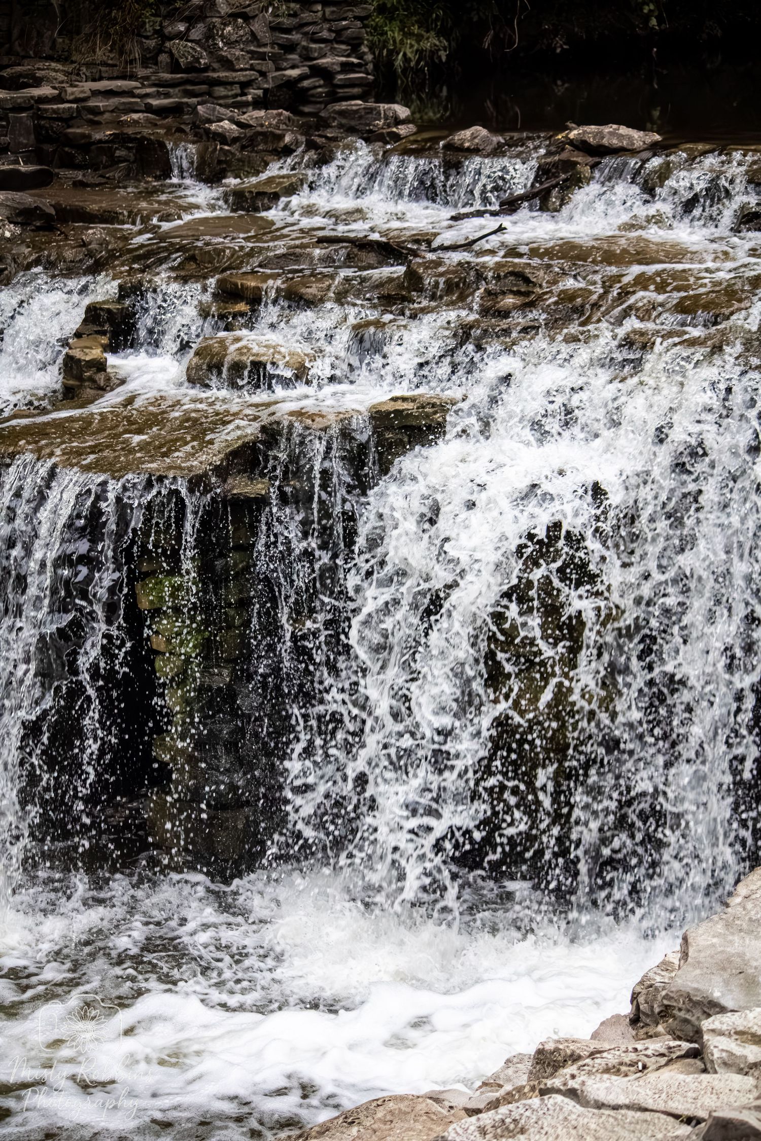 Cascading waterfall flows over rocky ledges creating multiple tiers of white rushing water against dark stone.