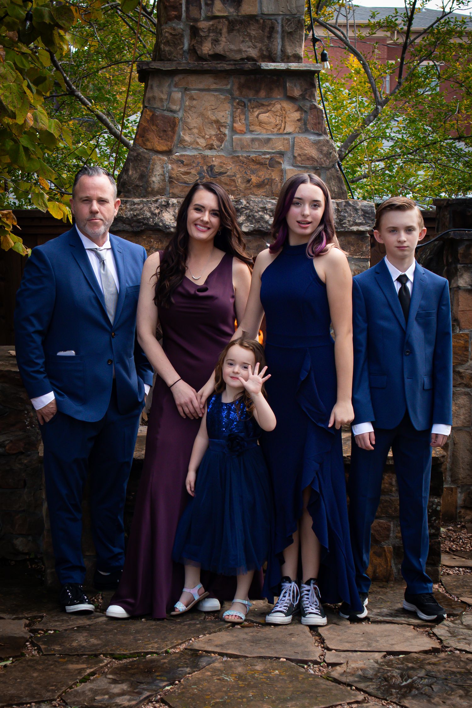 Family of five posing in coordinating blue and purple formal attire outdoors near a stone fireplace.