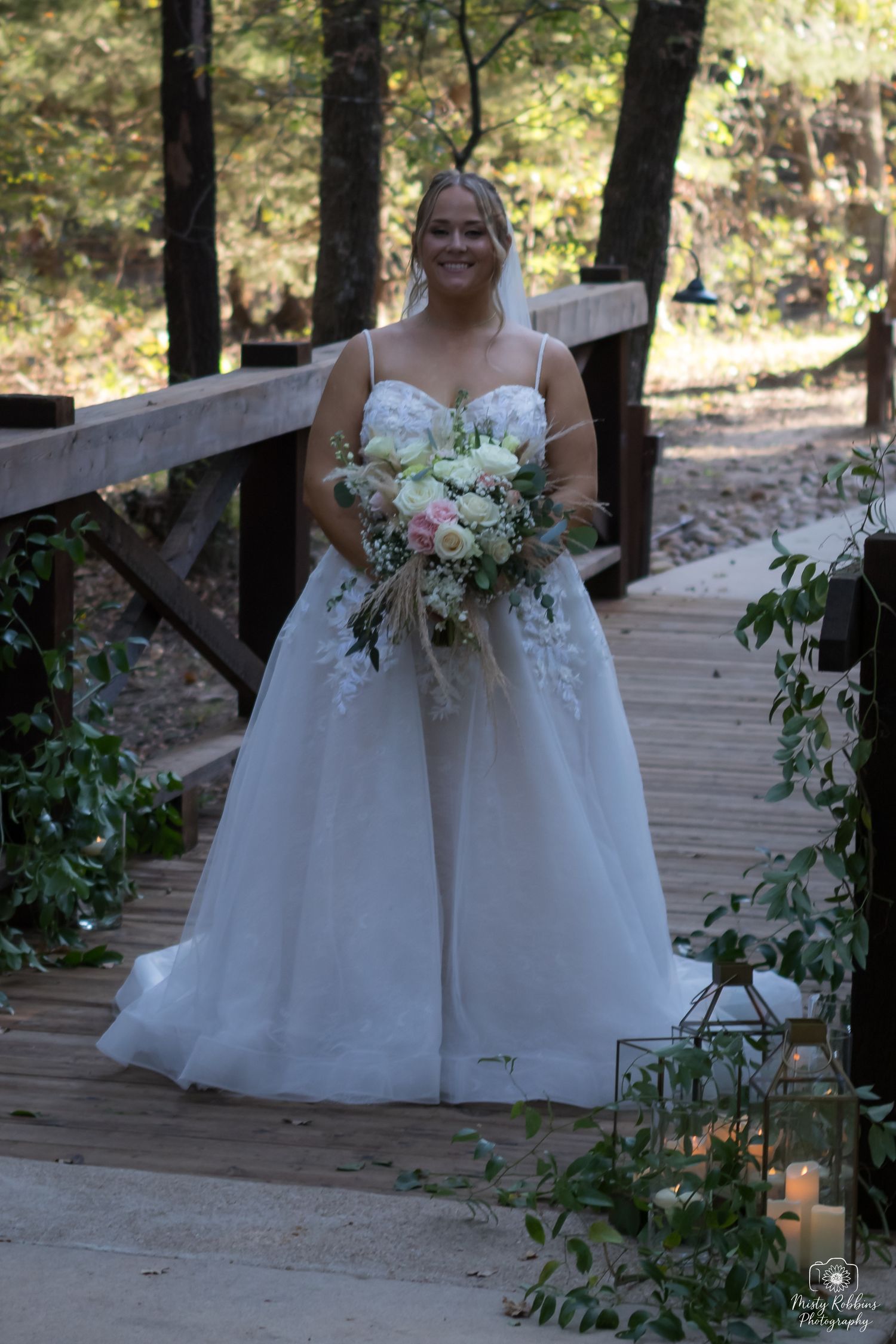 A bride in a white wedding gown walks along a wooden boardwalk surrounded by greenery in a forest setting.
