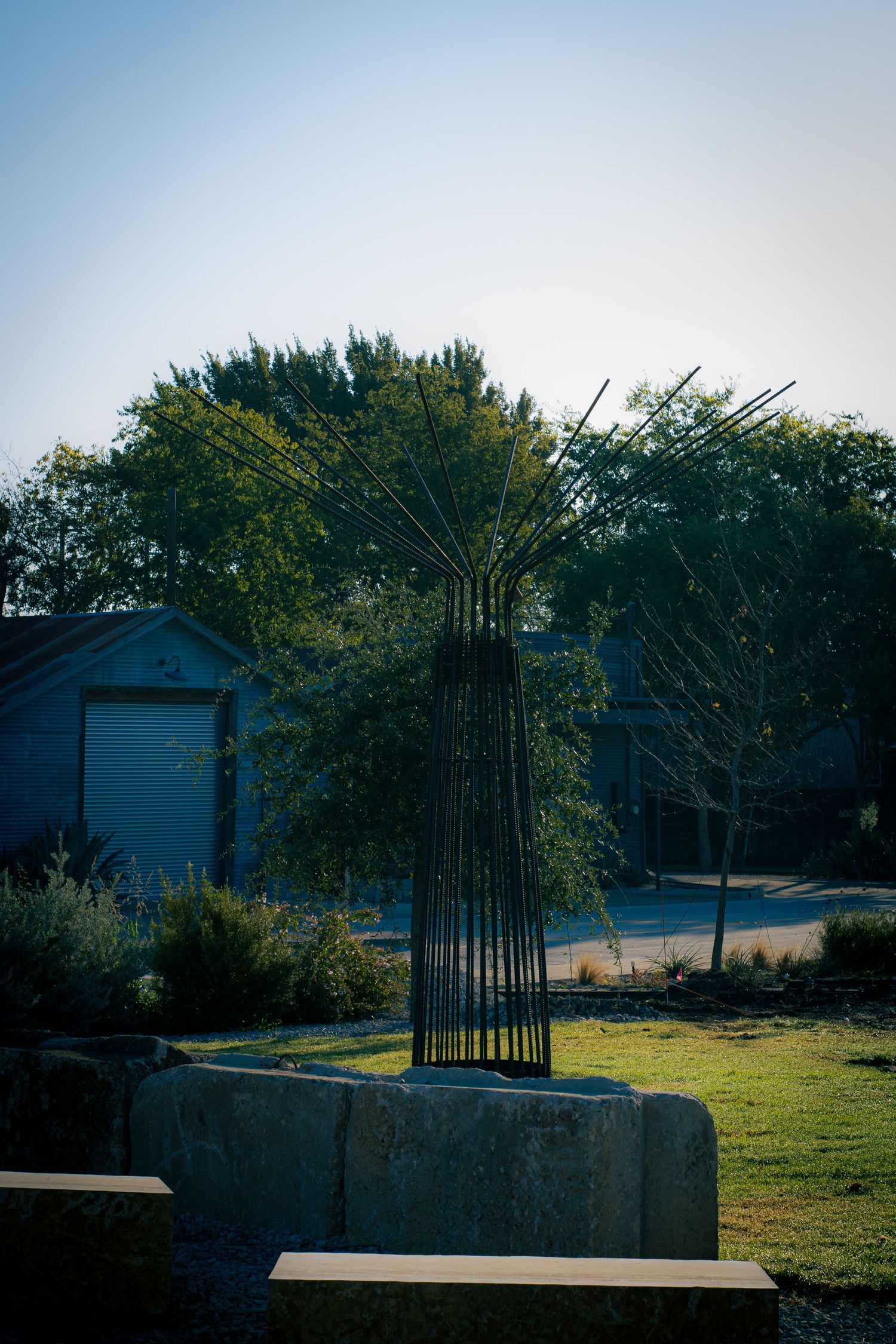 Modern metal sculpture rises from concrete base in garden setting with blue sheds in background.