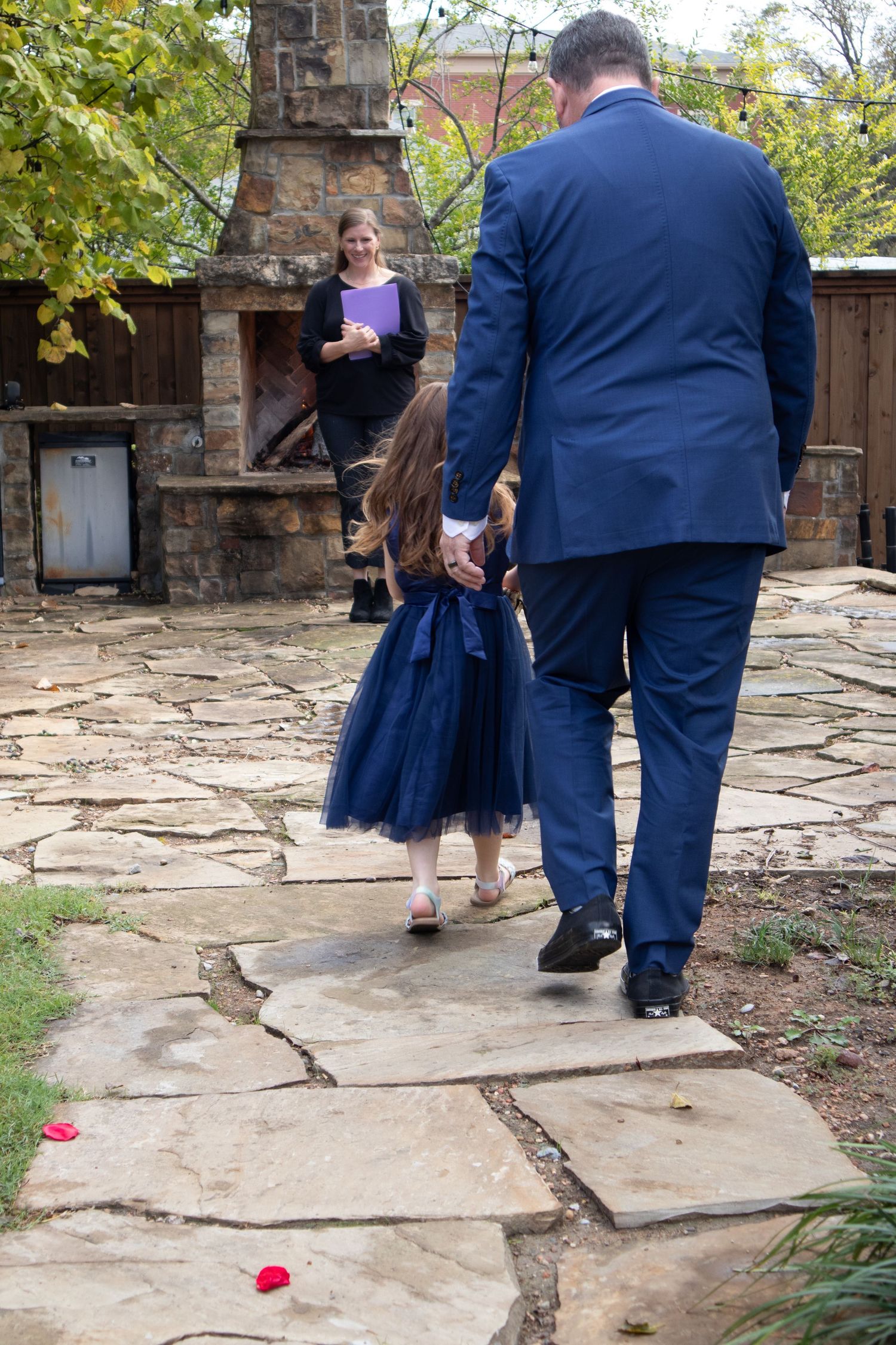 A person in a navy suit holds hands with a small child in a blue tulle dress on a stone patio.