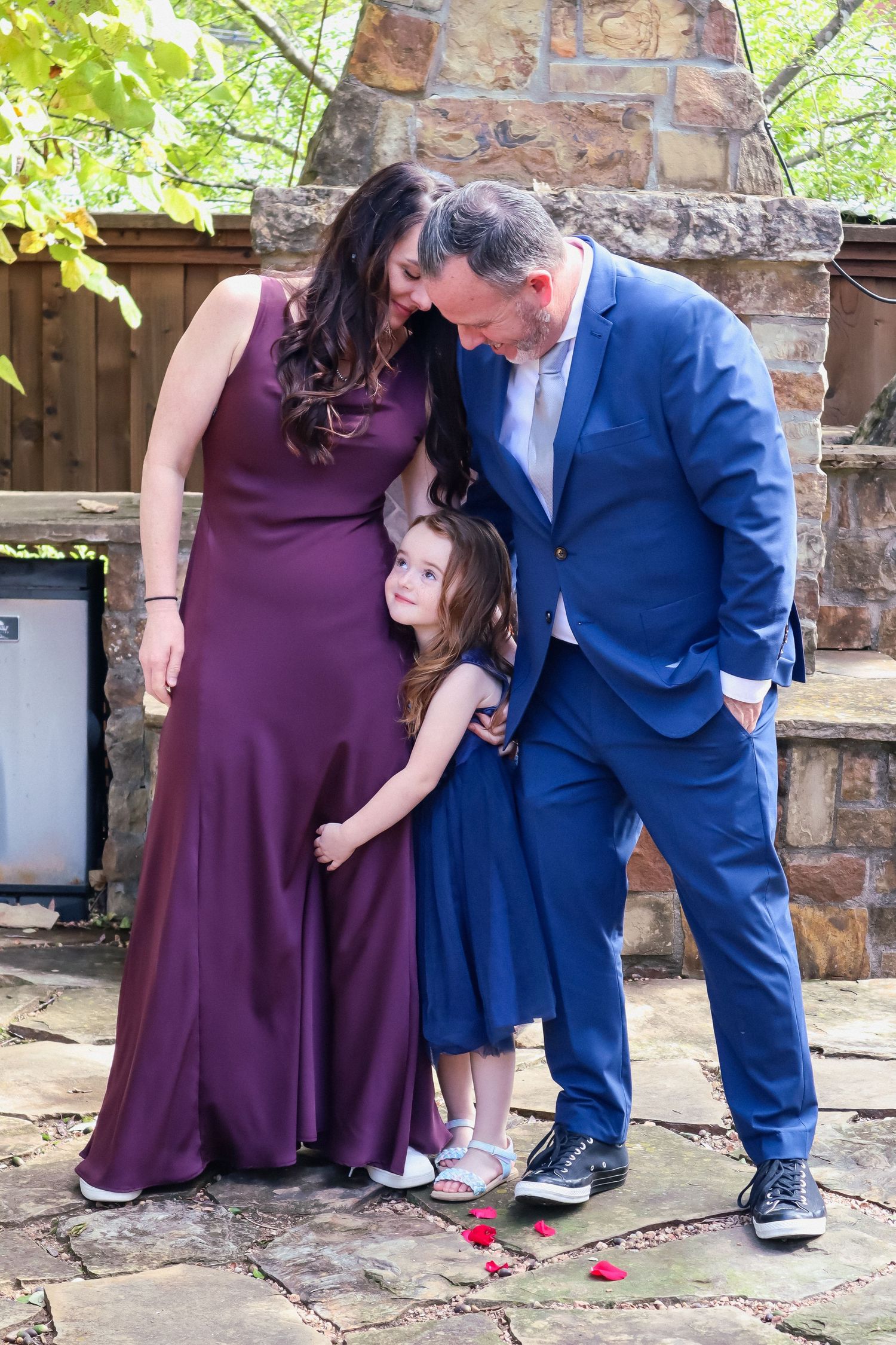 Three people in formal attire share an intimate moment on a stone patio near a fireplace.