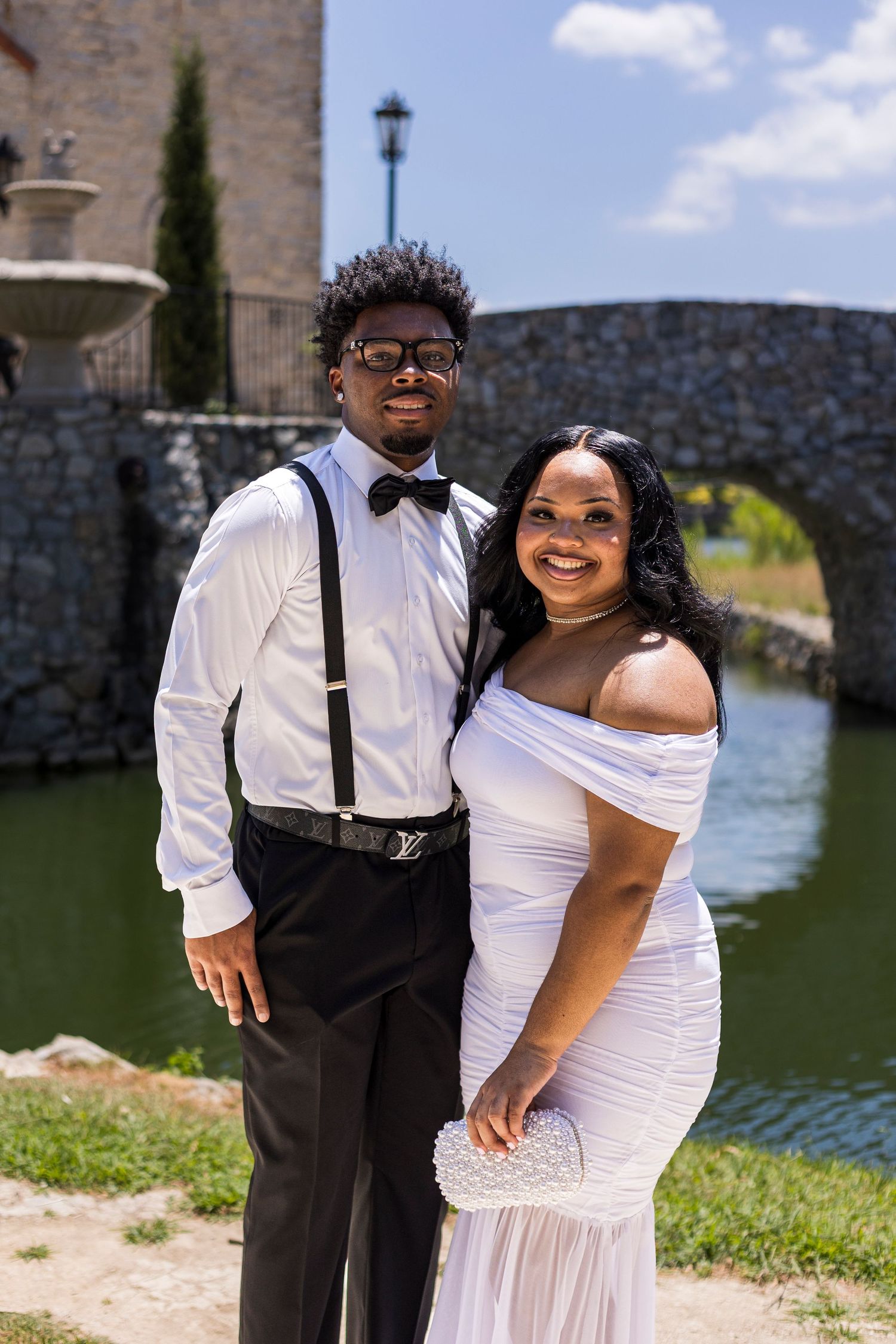 Couple posing outdoors near a stone bridge and fountain, man in suspenders and bow tie, woman in white off-shoulder gown.