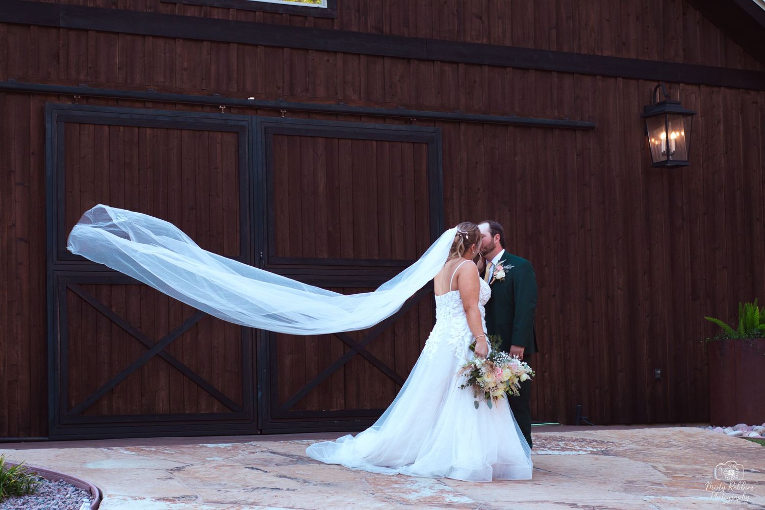 A flowing white wedding veil catches the breeze against a rustic wooden barn backdrop.