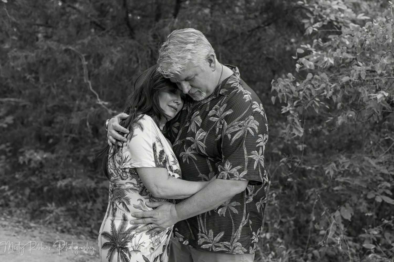 Black and white photo of a couple embracing tenderly outdoors, surrounded by lush trees in a romantic portrait.