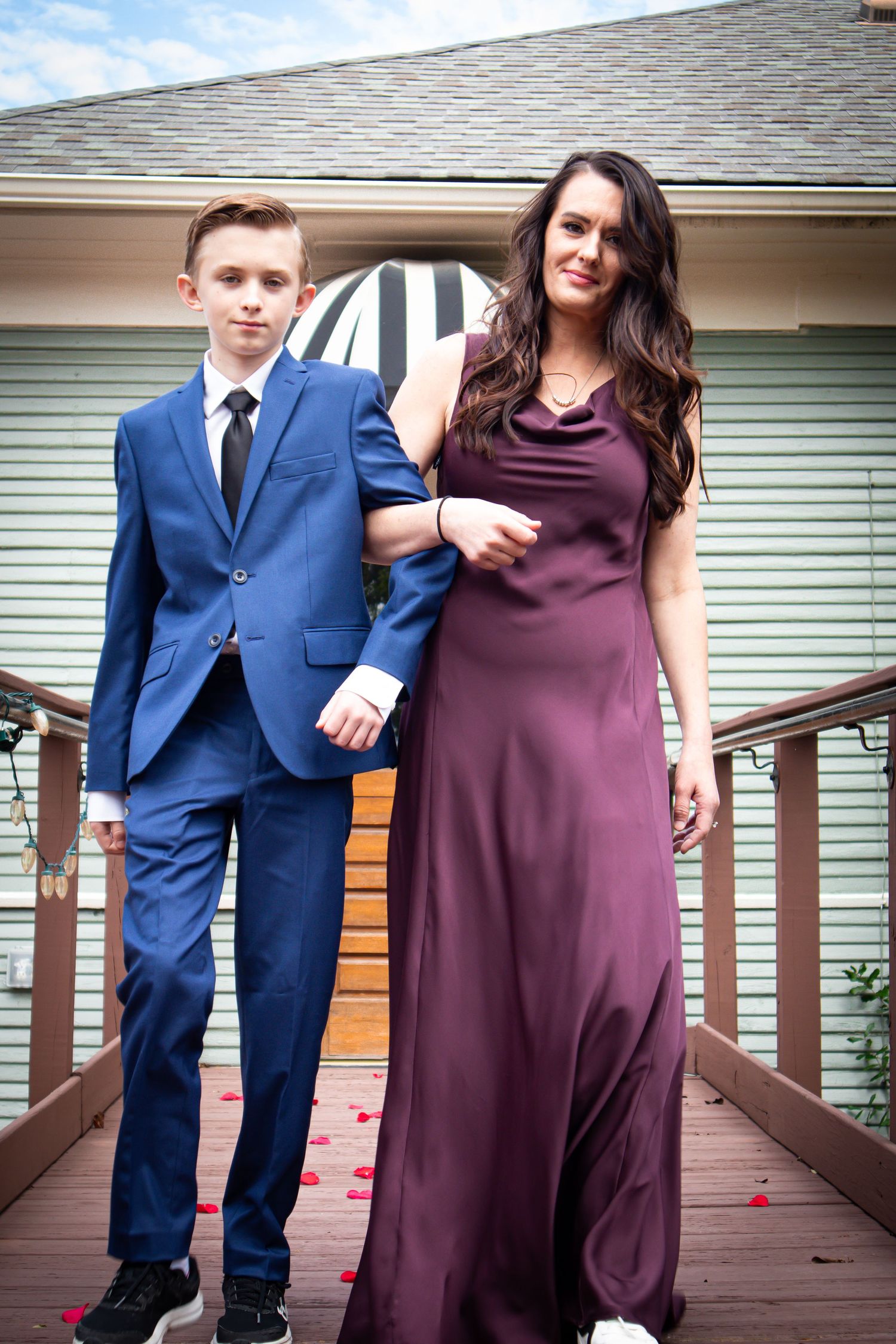 A young person in a blue suit and a woman in a burgundy gown pose together on a wooden deck.