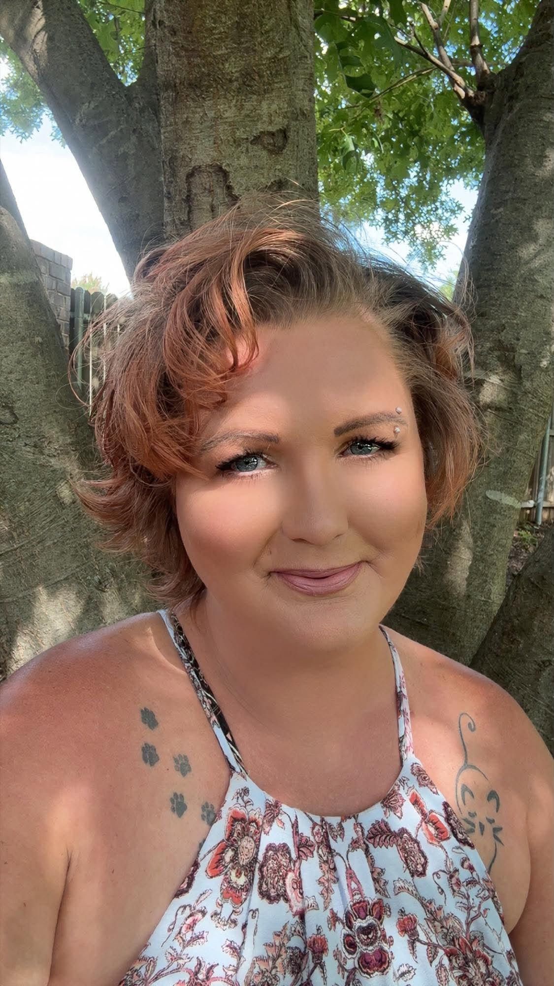 Woman with short auburn hair and floral tattoos smiles outdoors near a large tree on a sunny day.