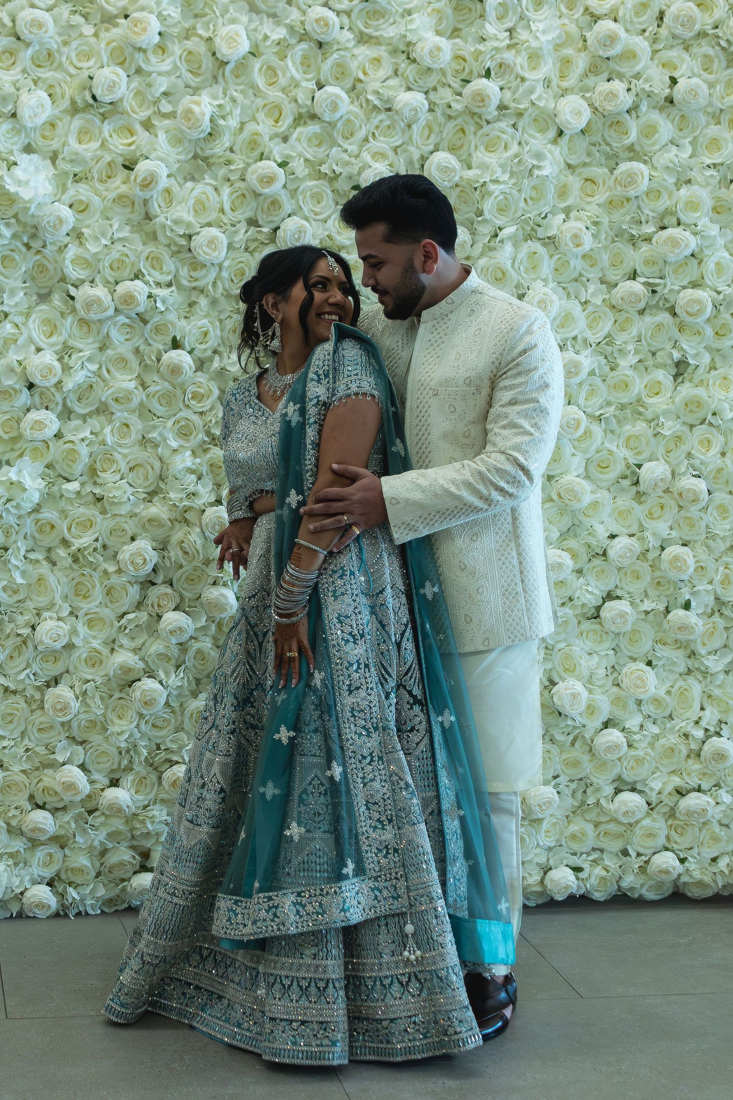 Indian couple in traditional wedding attire posing before a white floral wall, bride in teal lehenga, groom in cream sherwani.
