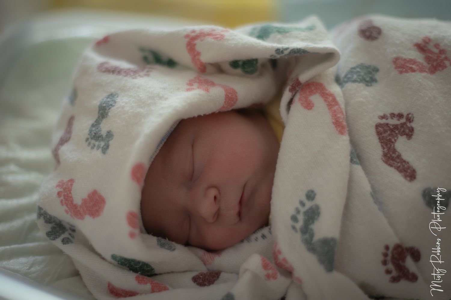 Newborn baby swaddled in a white blanket with colorful footprint patterns, sleeping peacefully in hospital.