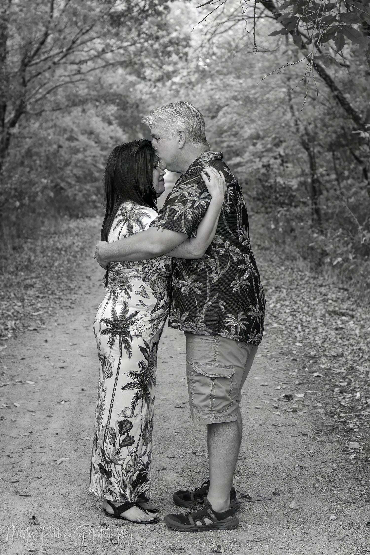 Black and white photo of a couple sharing a romantic kiss on a wooded trail, both wearing tropical print outfits.