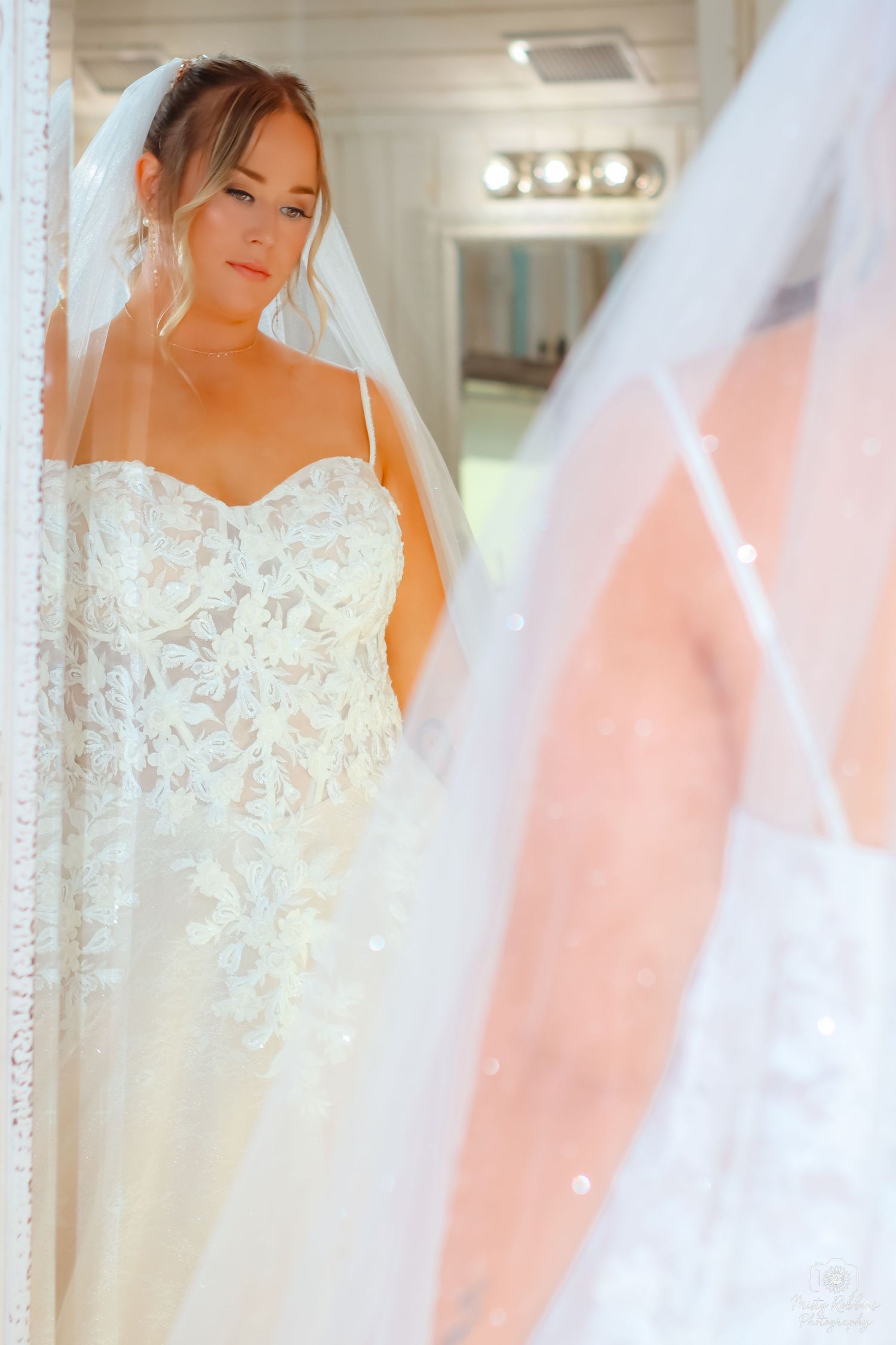 A bride in a delicate white lace wedding gown with a flowing veil stands elegantly in front of mirrors.