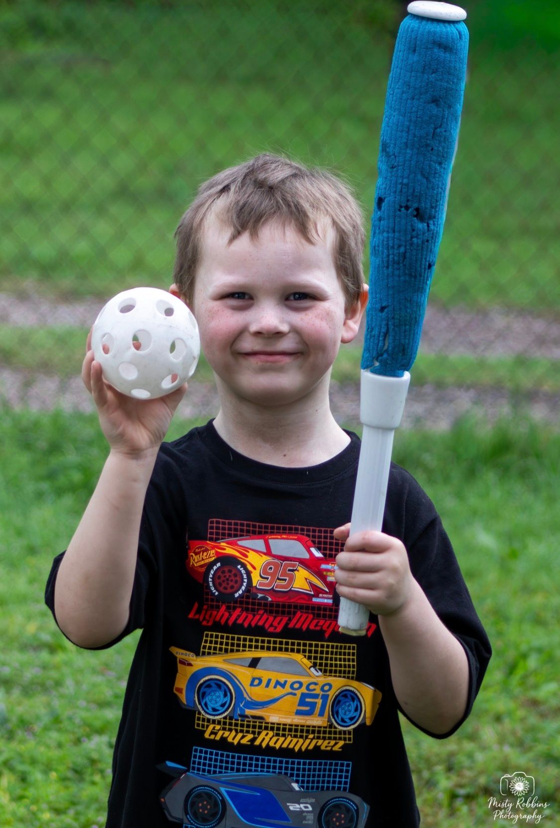 Young boy smiling outdoors holding a blue foam baseball bat and white wiffle ball, wearing a Cars Lightning McQueen shirt.