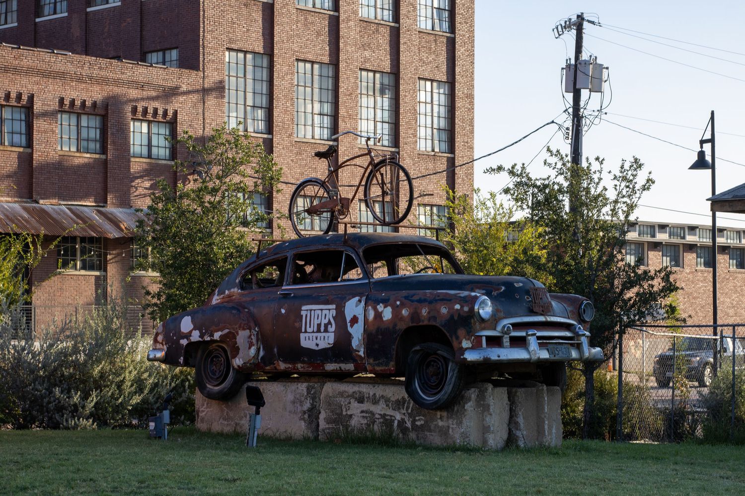 Rusty vintage car with bicycle on roof displayed as outdoor art sculpture near brick building.